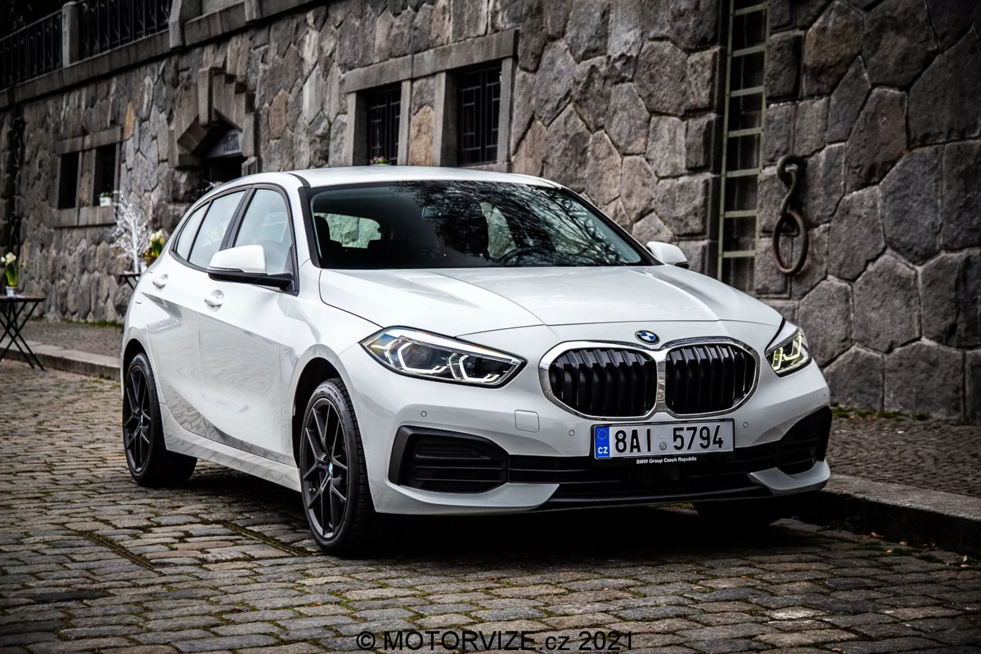 A imagem mostra um BMW Série 1 de 2019, especificamente o BMW 116i, em cor branca. Fotografado de um ângulo frontal de três quartos, mostra a frente do carro, o lado do passageiro e o design detalhado das suas rodas. A foto é tirada de um ponto de vista ligeiramente elevado, sugerindo que o fotógrafo estava em pé ou usando um tripé. O carro está estacionado com as rodas dianteiras viradas para a direita, revelando uma vista clara das jantes e do perfil lateral do pneu. As características incluem uma grade frontal proeminente com lâminas verticais, linhas esculpidas no capô e nos painéis laterais, faróis LED delgados e entradas de ar no para-choque que podem conter sensores de estacionamento. Os espelhos laterais com indicadores de direção integrados e as janelas sem moldura destacam a estética premium. Rodas liga leve de múltiplos raios sugerem um nível de acabamento superior ou uma atualização opcional. A janela traseira inclinada acentua o perfil do hatchback. O veículo está estacionado em uma calçada de paralelepípedos em um ambiente urbano ou histórico com um prédio de pedras e uma pequena mesa ao estilo bistrô ao fundo. A imagem é nítida e detalhada, capturada sob luz natural e nublada.
