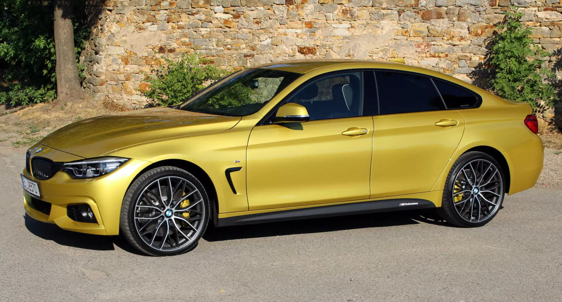 Coupé BMW Série 4 dourado metálico de 2013 estacionado paralelamente a um antigo muro de pedra, exibindo seu perfil lateral aerodinâmico e elegante, rodas de liga leve e melhorias tecnológicas de um ângulo levemente elevado