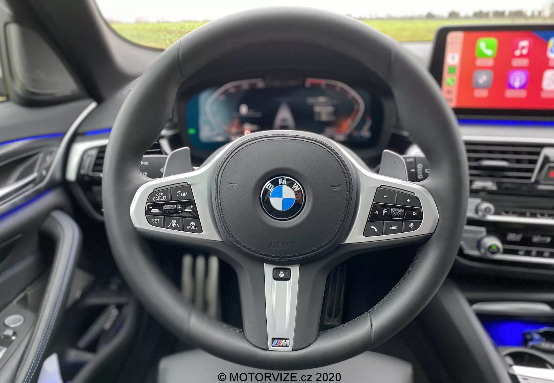 Vista interior da perspectiva do condutor dentro de um BMW 520i Touring Steptronic 2020, destacando um volante esportivo de três raios com emblemas BMW e M. O volante possui vários botões de controle para controle de cruzeiro, mídia, comando de voz e funções de telefonia, e paddle shifters que indicam uma transmissão automática com opções manuais. Atrás do volante, um display totalmente digital mostra velocímetro, tacômetro e informações do veículo. À direita, é visível parte da consola central com uma grande tela de infotainment exibindo Apple CarPlay, enfatizando o luxo, tecnologia avançada e design orientado para o condutor.