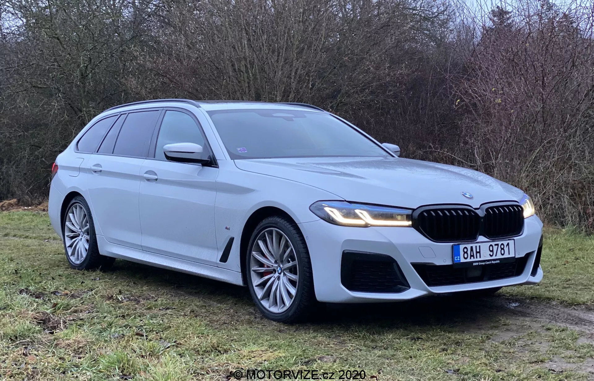 BMW 5 Series Touring 2020 prata estacionado na grama, visto de um ângulo frontal de três quartos, mostrando a grade frontal, faróis e design aerodinâmico. Possui faróis de LED, grandes rodas de liga, grade em forma de rim e sensores de estacionamento. Limpo e novo, destacando o emblema da BMW, setas integradas, trilhos de teto e antena tipo barbatana de tubarão.
