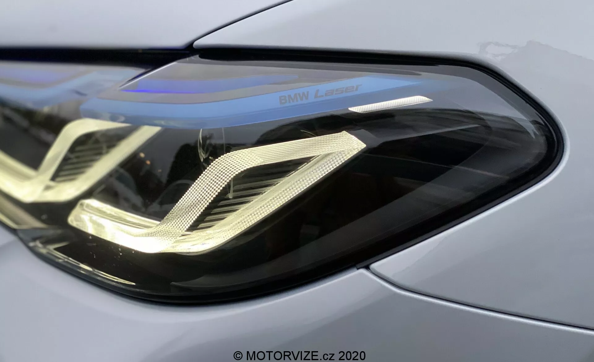 Close-up do farol dianteiro esquerdo do BMW 520i Touring Steptronic 2020, apresentando lente de policarbonato curva e elegante, luzes diurnas LED características em forma de dupla hélice, carcaça refletora e marca 'BMW Laser', destacando a avançada tecnologia de faróis a laser e painel de carroceria metálico premium.