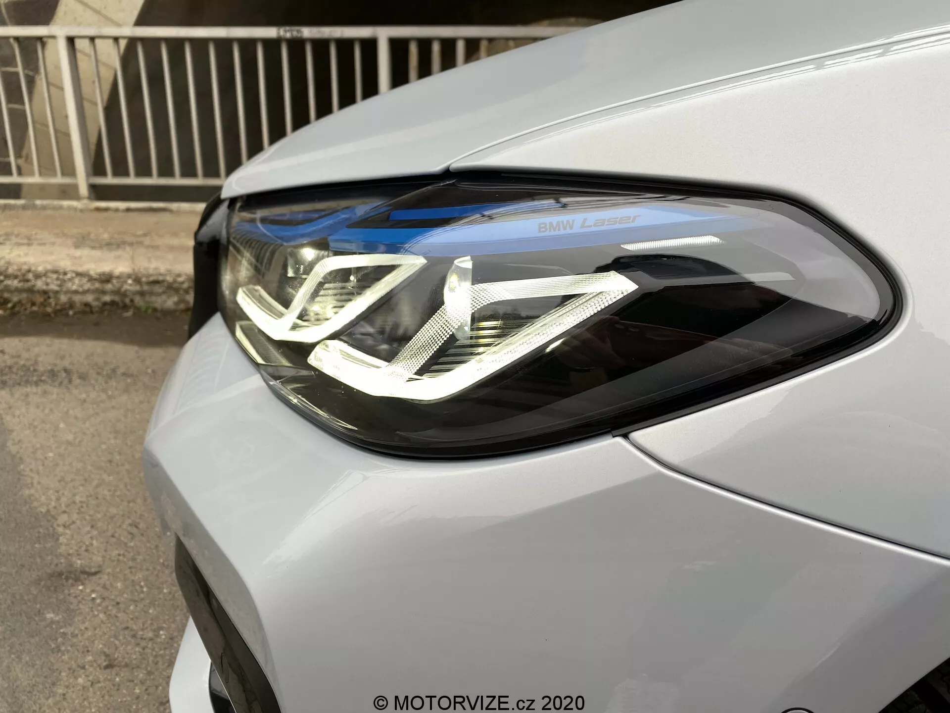 Close-up do farol do lado do motorista do BMW Série 5 2020 (BMW 520i Touring Steptronic). A imagem captura o carro sob um ângulo frontal de três quartos, destacando as intrincadas luzes diurnas de LED com design em forma de U duplo, lentes de projetor e inscrição 'BMW Laser'. A unidade do farol integra-se perfeitamente ao acabamento metálico ou perolado do carro.