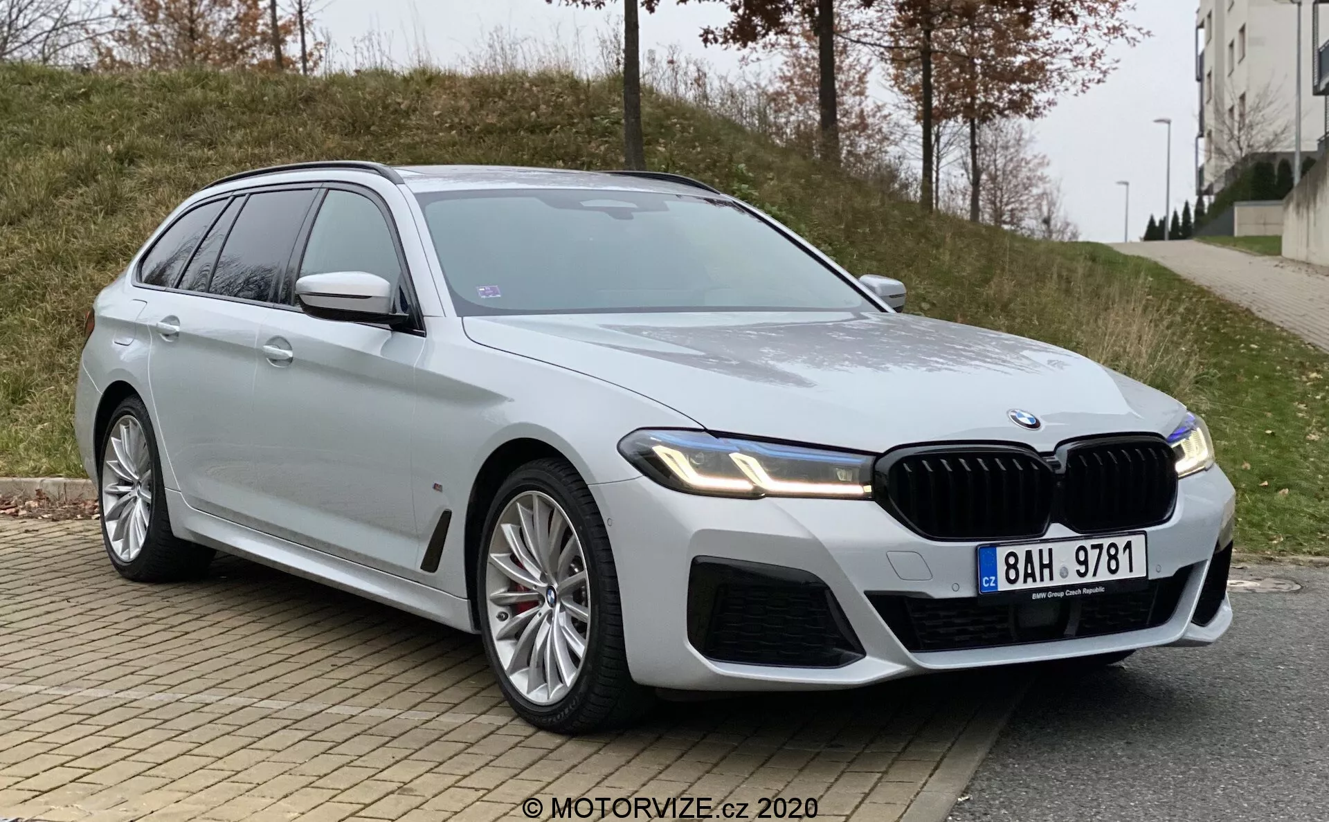 Um moderno e luxuoso BMW Série 5 (BMW 520i Touring Steptronic) station wagon fotografado a partir de um ângulo baixo de três quartos da frente. A imagem destaca a grade frontal do carro, os faróis angulares e elegantes provavelmente com tecnologia LED, e as linhas de design musculosas no capô e nos para-lamas. As partes visíveis incluem a grade frontal, para-choque, faróis, porta do motorista frontal, para-lamas dianteiro, parte da porta traseira, vidros laterais, rodas e espelho lateral do lado do motorista. As grandes rodas de liga sugerem uma variante mais esportiva do modelo, e a pintura prateada é reflexiva e limpa. O carro possui uma grade frontal proeminente com ripas verticais, um emblema no para-lamas dianteiro indicando um nível de acabamento superior e espelhos laterais com sinais de volta integrados. Um teto solar também é visível. O veículo está estacionado em uma estrada urbana com um meio-fio e calçada em primeiro plano e uma área gramada e um edifício ao fundo, sob condições de iluminação nublada.