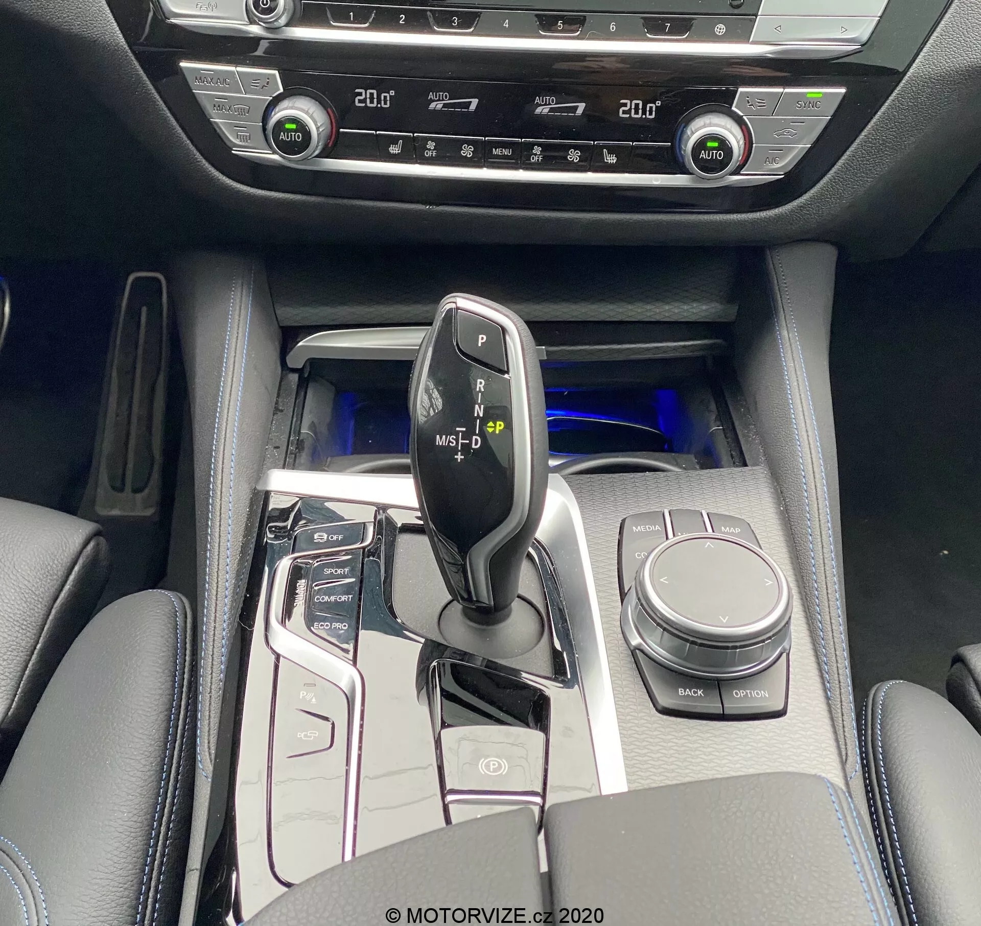 Vista de cima da consola central do BMW 520i Touring Steptronic 2020, capturando uma alavanca de câmbio preta brilhante com detalhes metálicos, cercada por um painel de controle de alto brilho com botões para sistema start/stop, modos de condução, sensores de estacionamento e mais. Visíveis são o controle rotativo iDrive, porta-copos e apoio de braço acolchoado com armazenamento. A iluminação ambiente azul fria aumenta a sensação de luxo. Controles de climatização dual-zone e aquecimento de assentos indicam funções de conforto avançadas. Os materiais interiores incluem superfícies semelhantes ao couro com detalhes metálicos e painéis de alto brilho.