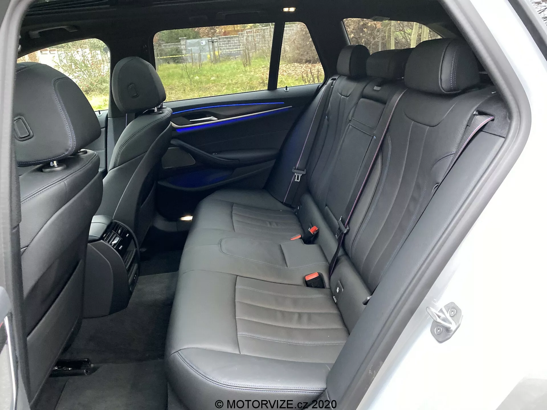Interior do BMW 520i Touring Steptronic 2020 focando na área dos assentos traseiros do lado direito. O ponto de visão está próximo à porta traseira direita do passageiro, mostrando um banco para três pessoas com estofamento de couro escuro e costura decorativa. As características incluem três apoios de cabeça, três cintos de segurança, um painel de porta com uma grade de alto-falante, alça e elementos de controle, uma faixa de iluminação ambiente azul e uma rede de armazenamento na parte de trás do banco do passageiro dianteiro. O interior bem conservado não possui pertences pessoais visíveis ou desgaste significativo. A vista parcial externa através da janela traseira inclui uma cerca.