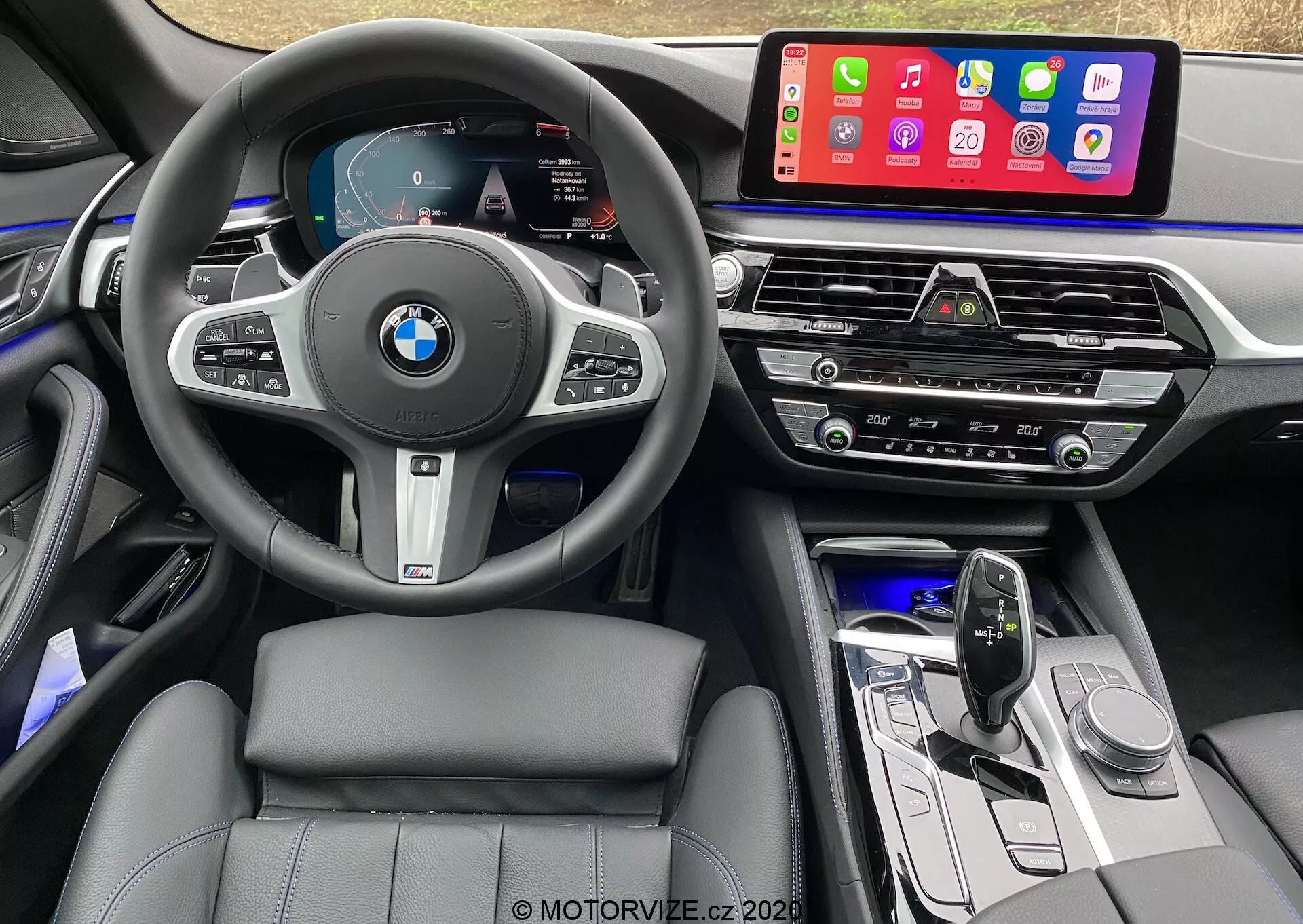 Vista interior do banco do motorista de um BMW 520i Touring Steptronic 2020, com um volante multifuncional revestido em couro com o logotipo da BMW e botões de controle para controle de cruzeiro, áudio e telefonia. O painel de instrumentos digital exibe velocímetro, tacômetro e um mapa de navegação. O console central abriga um grande sistema de infoentretenimento com tela sensível ao toque que mostra aplicativos móveis como Apple CarPlay. Abaixo estão saídas de ar, painel de controle de clima, controles de áudio, alavanca de câmbio de design elegante e botões de função. O interior apresenta assentos com costura de couro, iluminação ambiente e materiais de alta qualidade com acentos metálicos.
