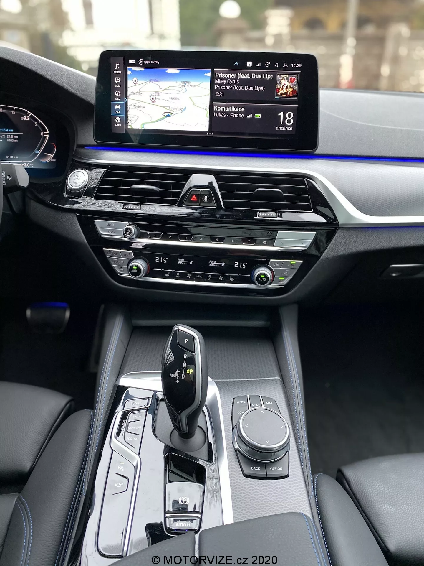 Vista interior do BMW Série 5 2020 (BMW 520i Touring Steptronic), mostrando a consola central e o painel de instrumentos da perspectiva do passageiro dianteiro. A consola central apresenta um seletor de marchas automático elegante em modo 'park', com vários controles incluindo um botão rotativo para o sistema de infotainment e botões para modos de condução ou configurações do veículo. A seção central do painel de instrumentos inclui controles de clima com botões e dials intuitivos, acima dos quais há uma slot para CD. O topo do painel de instrumentos apresenta proeminentemente uma tela sensível ao toque de infotainment larga com mapa de navegação e informação de limite de velocidade. Parcialmente visível está o cluster de instrumentos digitais mostrando o velocímetro. Os materiais internos destacam acabamentos premium, contrastando couro escuro com uma faixa de acento azul ao longo de toda a largura do painel de instrumentos.