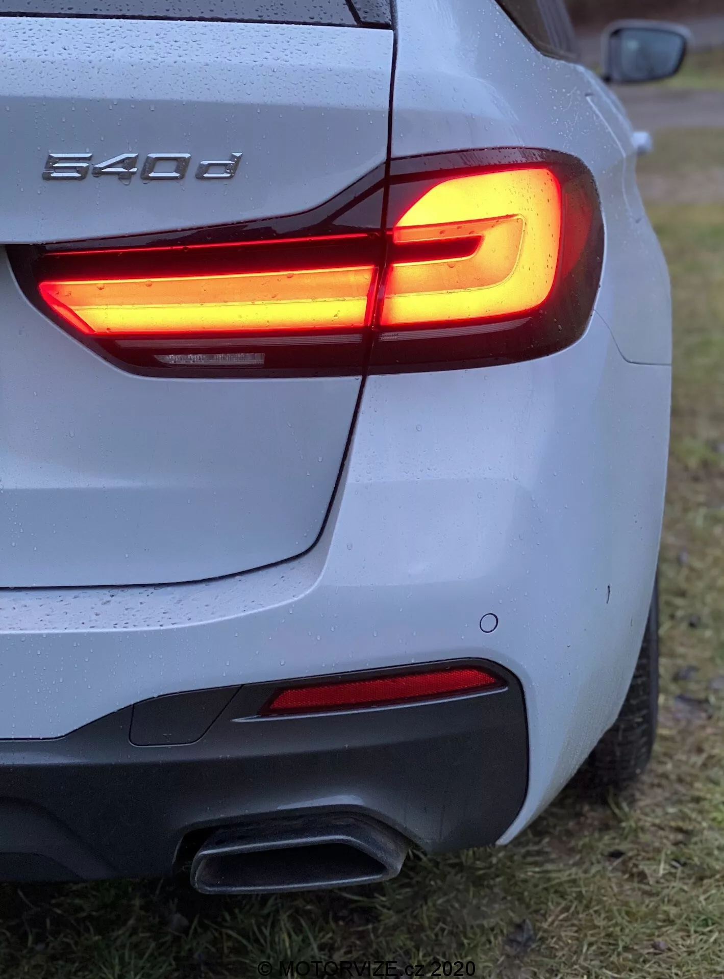 Vista de ângulo traseiro três quartos de um BMW 540i, exibindo luz traseira LED em forma de L, tubos de escape duplos, carroçaria prateada/cinza com gotas de água, para-choque traseiro esculpido, emblema parcial da BMW, estacionado em área gramada.