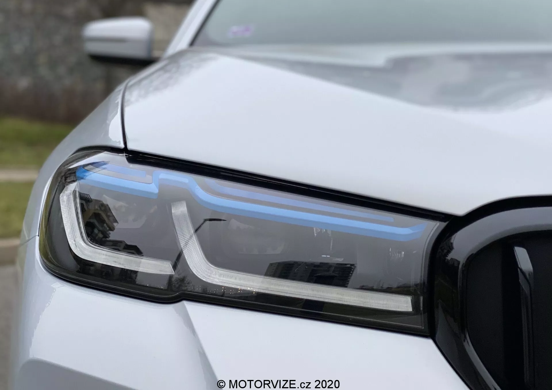 Vista aproximada do conjunto de farol do lado do motorista do BMW 520i Touring Steptronic, com foco nas luzes diurnas LED, farol projetor e indicador de direção. A fotografia mostra a seção frontal esquerda do carro, incluindo parte do capô, para-choque dianteiro e para-lama, com a grade visível à direita. A foto apresenta uma profundidade de campo rasa que destaca a tecnologia e o design do farol em luz natural difusa com acabamento de pintura branca não metálica.