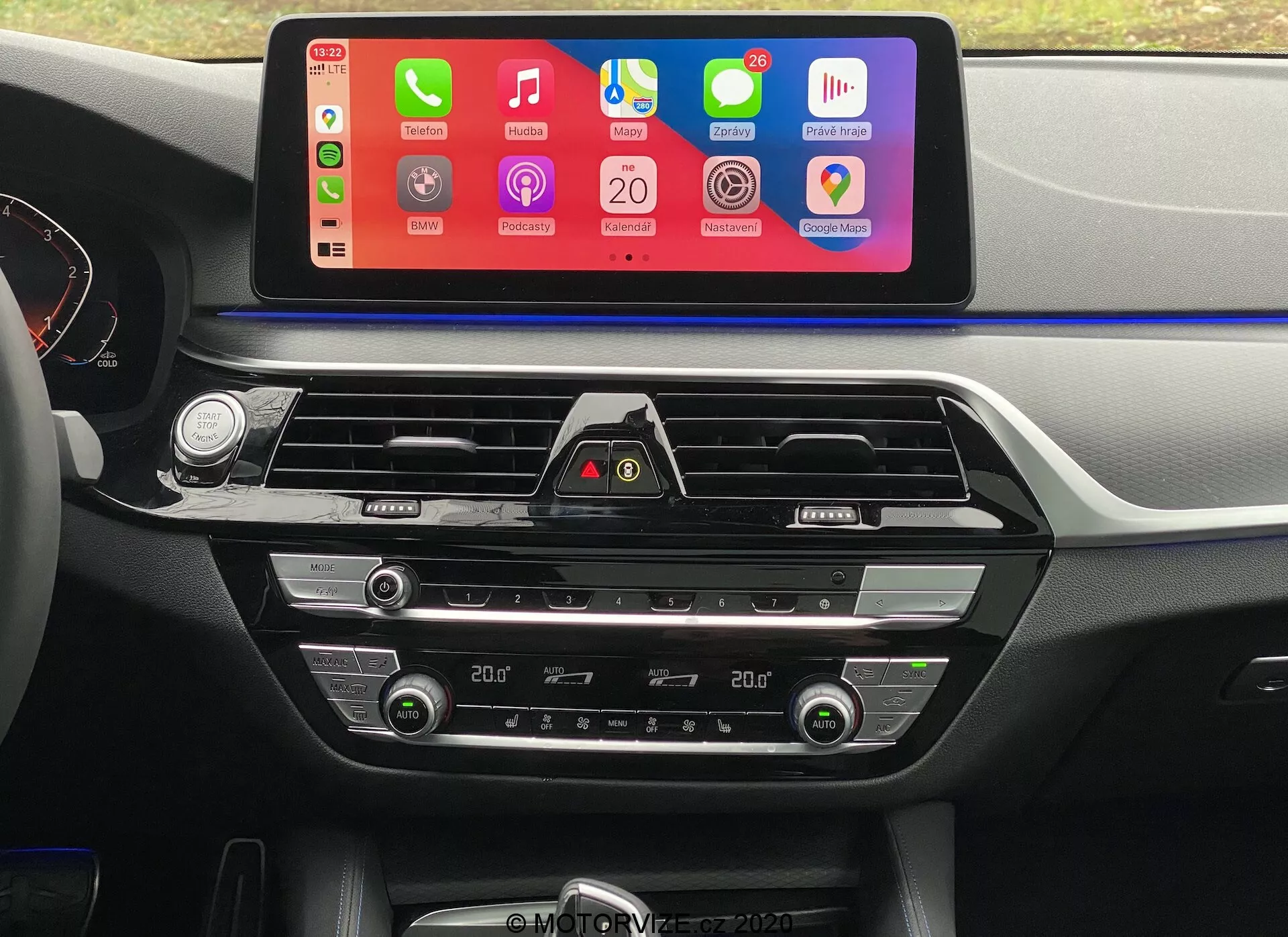 Imagem interior de um BMW 5er-Reihe 2020 (BMW 520i Touring Steptronic) na perspetiva do assento do condutor, focando-se no painel central. A característica mais proeminente é o grande ecrã tátil de infoentretenimento orientado horizontalmente, com uma interface colorida que suporta a integração de smartphones, como Apple CarPlay e Android Auto. Abaixo do ecrã, são visíveis saídas de ar com detalhes cromados e um botão de luzes de emergência. Mais abaixo, encontram-se os controlos de climatização de duas zonas com ecrãs digitais para as definições de temperatura, botões e discos para ajuste da temperatura do ar, velocidade do ventilador e distribuição do ar. O painel de instrumentos e a consola central são feitos de uma combinação de plásticos suaves ao tato e detalhes metálicos, aumentando o apelo estético.