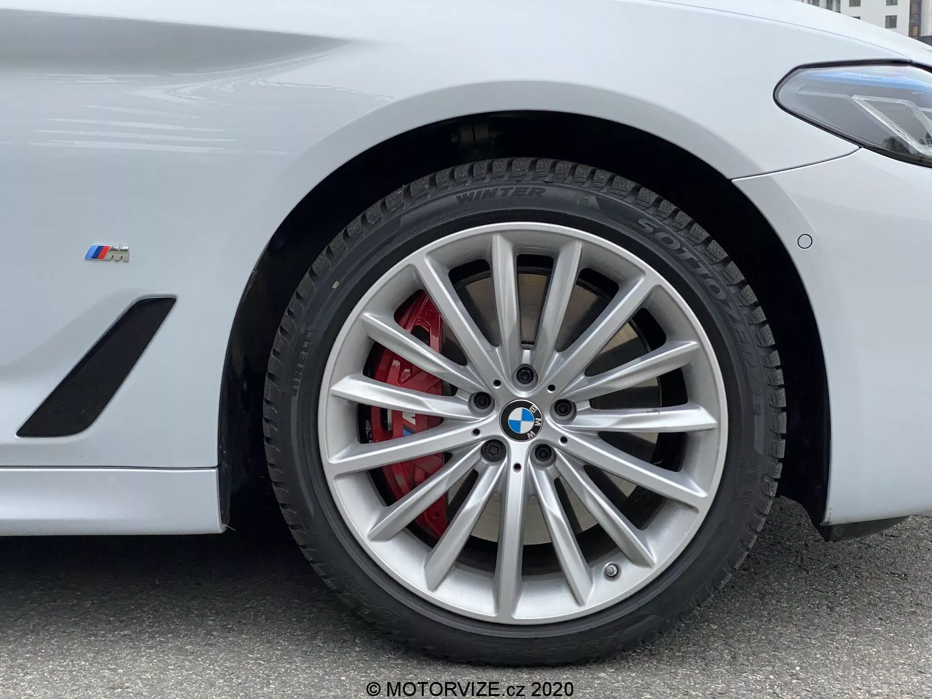 Imagem em close-up da seção da roda dianteira de um BMW da Série 5 2020, especificamente o modelo 520i Touring Steptronic. A fotografia em ângulo baixo destaca o intrincado design da roda de liga leve com múltiplos raios e acabamento prateado, complementada por uma pinça de freio vermelha e um rotor ventilado. O emblema da BMW e o emblema 'M' no para-lama indicam que este é um veículo focado em desempenho da série M da BMW. A imagem captura detalhes-chave adequados para mostrar sistemas de freios de alto desempenho, elementos de design de veículos de luxo e tecnologia de pneus.