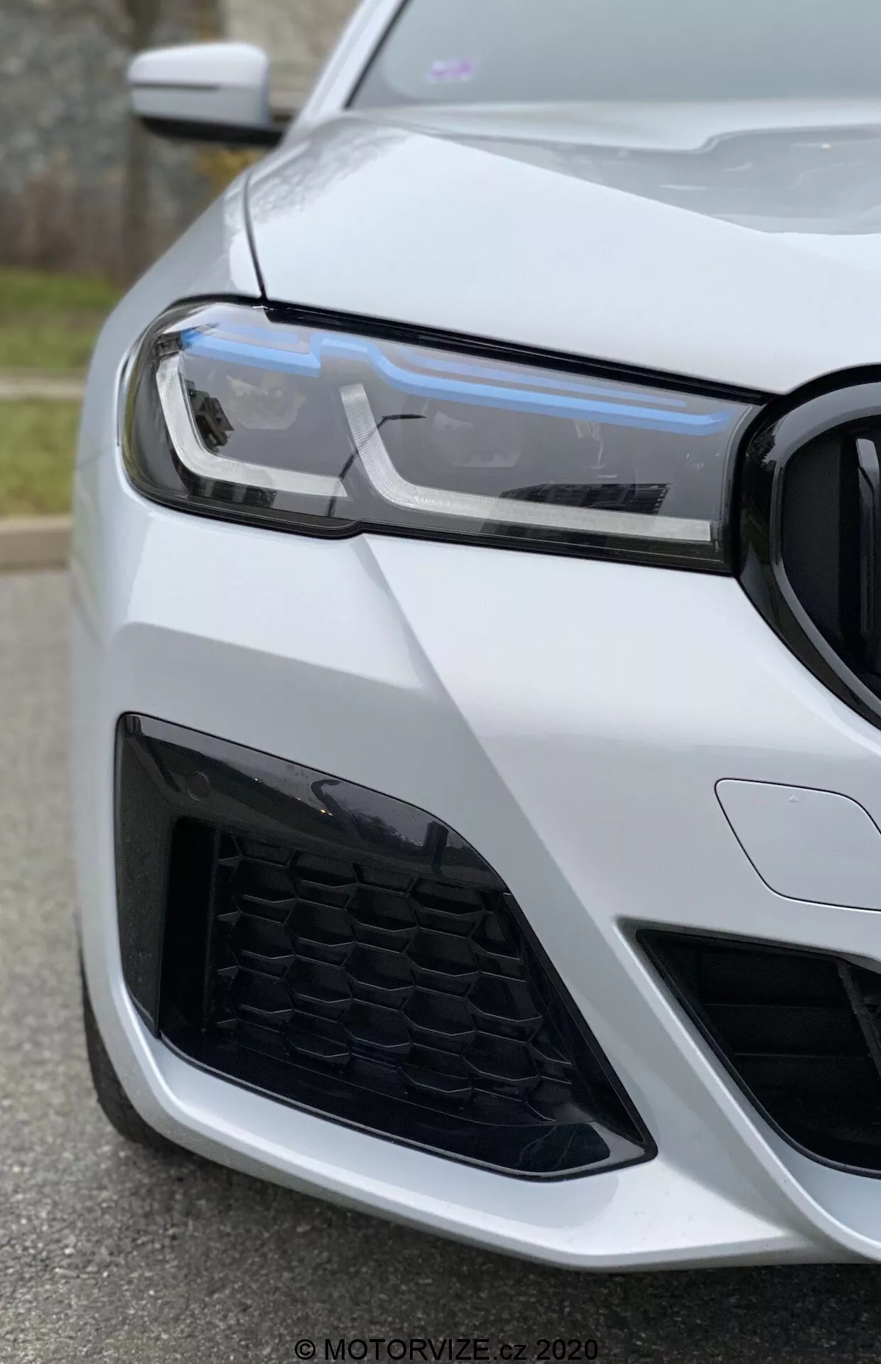 Vista em close-up da parte frontal esquerda de um BMW 520i Touring Steptronic (2020) tirada de um ângulo baixo, focando no farol, na grelha e no para-choques. As luzes diurnas LED modernas, o padrão em favo de mel da grelha e o design agressivo da entrada de ar sugerem um veículo esportivo e avançado. O carro é pintado de branco brilhante, destacando sua aparência luxuosa e bem cuidada.