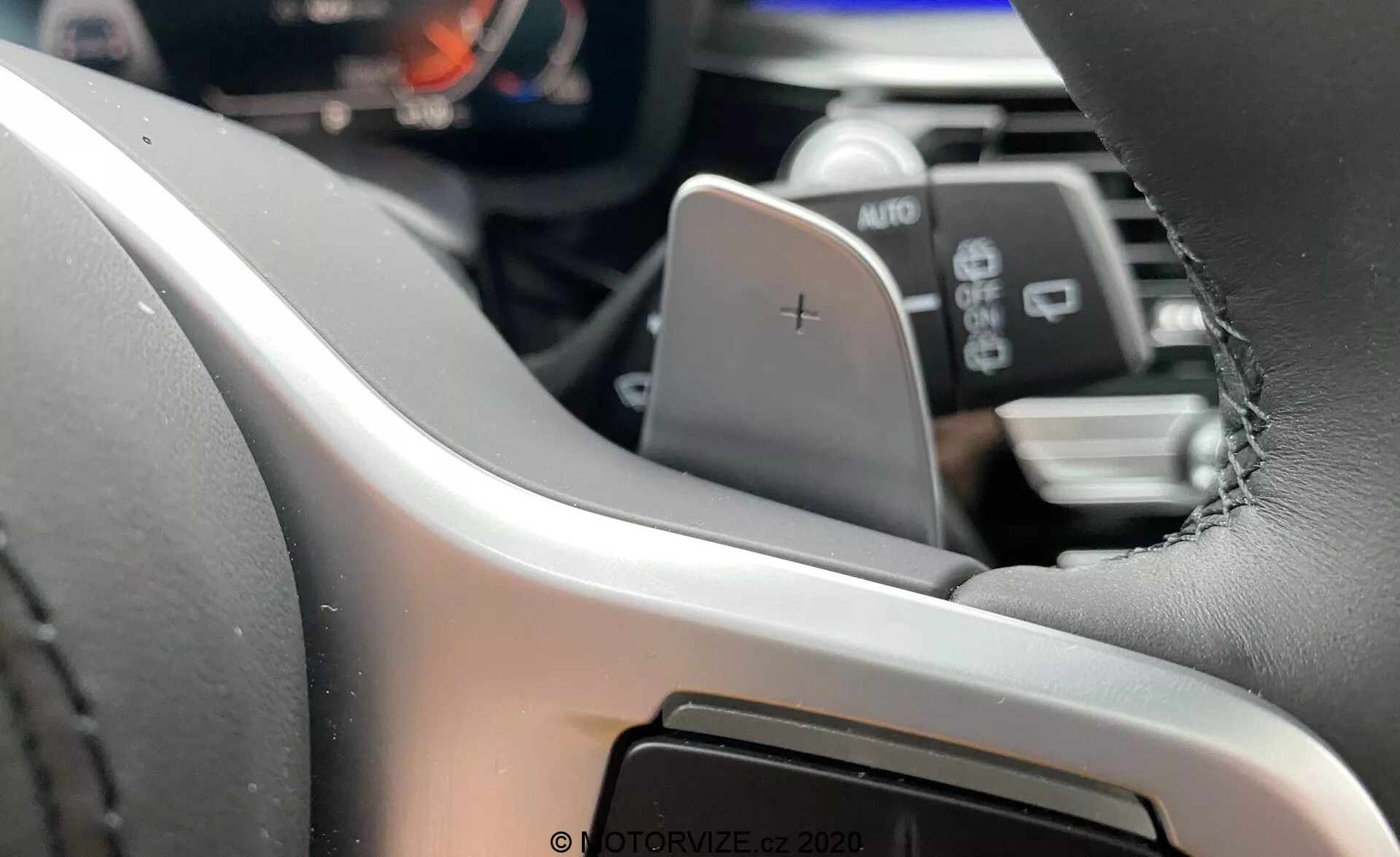 Vista interior do BMW Série 5 2020 (BMW 520i Touring Steptronic) focada no lado esquerdo do volante a partir da perspetiva do condutor⁠—volante moderno revestido a pele com costuras visíveis, equipado com uma pá para aumento de marchas marcada com um '+'. O painel de instrumentos digital ou cluster de instrumentos é parcialmente visível, destacando sistemas avançados de informação para o condutor. Abaixo da pá, uma alavanca rotulada AUTO indica provavelmente os controlos dos faróis ou dos limpa-para-brisas. No fundo desfocado, vislumbra-se o sistema de infoentretenimento ou consola central, sugerindo uma interface de ecrã tátil.