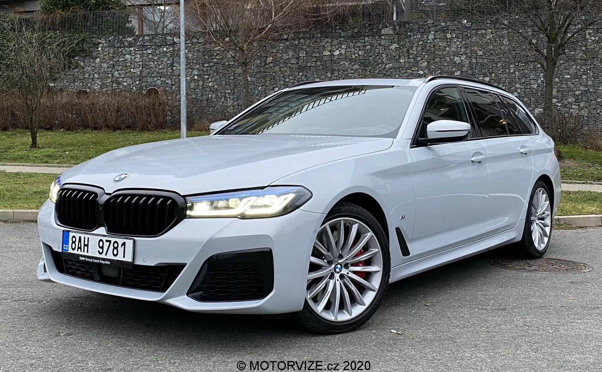 Veículo de luxo BMW Série 5 520i Touring Steptronic 2020 em luz natural diurna, apresentando a icônica grade de rim da BMW com faróis LED, parachoque dianteiro proeminente com entradas de ar, grandes rodas de liga leve multirraiadas, espelhos laterais montados nas portas com sinais de giro integrados, perfil aerodinâmico de estilo perua, linhas de caráter nas portas laterais, vidros traseiros escurecidos e freios a disco visíveis, em um cenário externo com uma estrada pavimentada, meio-fio gramado e uma parede de pedra.