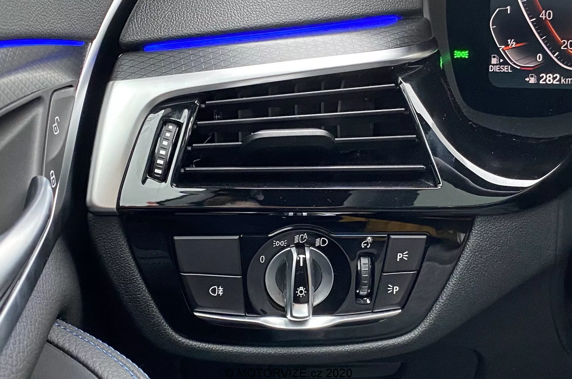 Close-up do interior do BMW Série 5 2020, focando no painel do lado do motorista com faixa de iluminação ambiente azul acima da ventilação horizontal. Os controles centrais incluem um dial rotativo para configurações automáticas e de faróis, botões provavelmente para luzes de neblina e nivelamento de faróis, botões adicionais de assistência ao motorista e um velocímetro digital mostrando 0 km/h com o veículo em modo Park e leitura de 282 km. Um indicador 'DIESEL' também é visível.