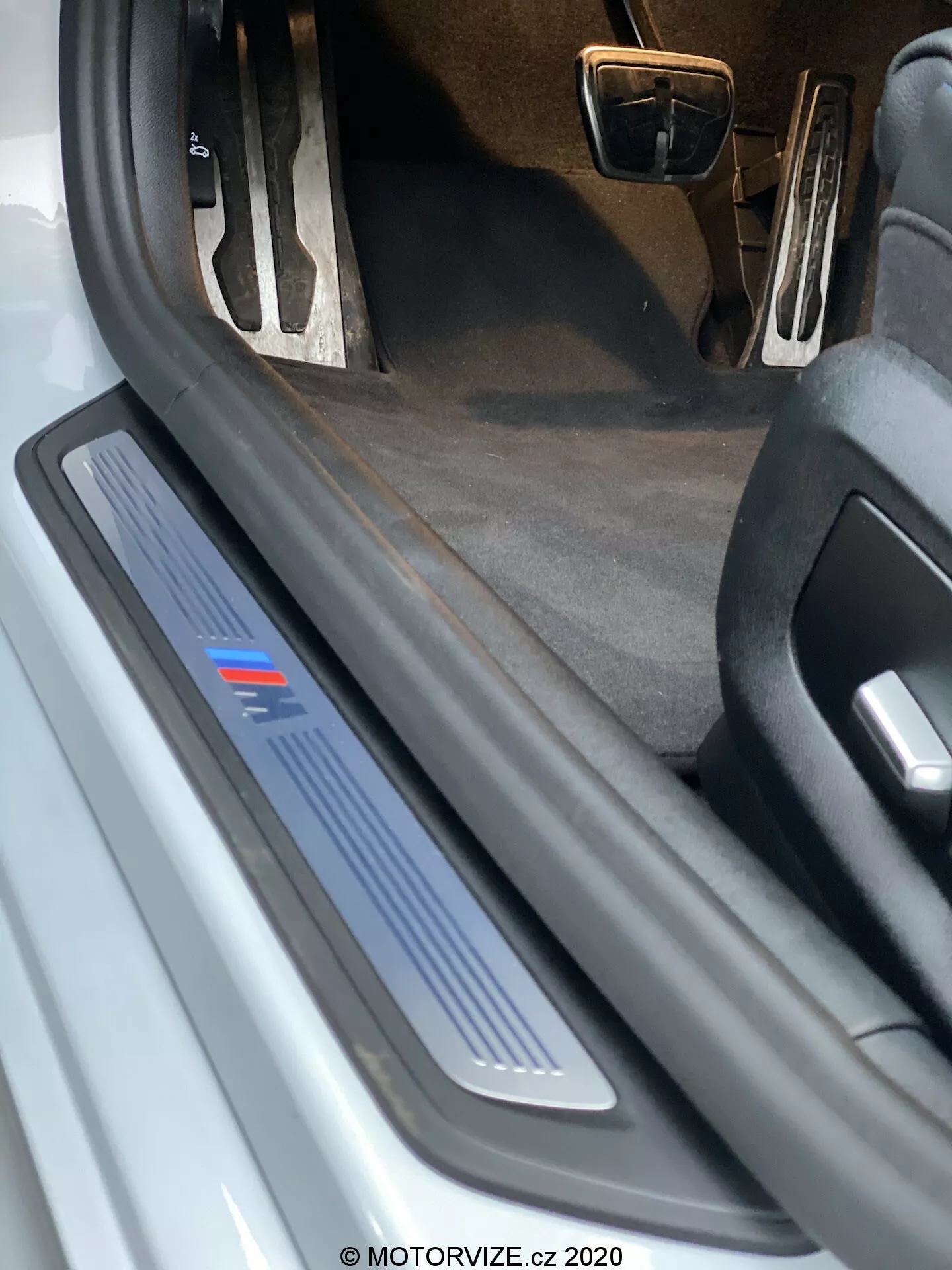 Vista aproximada do batente da porta do lado do motorista de um BMW 520i Touring Steptronic 2020. A imagem mostra uma perspectiva de ângulo baixo, destacando a placa de batente de metal escovado com uma faixa azul, que pode indicar um nível de acabamento específico ou edição esportiva. A porta aberta não é visível, mas a placa de batente protetora e decorativa é evidente, sugerindo possível iluminação LED. O interior escuro do carpete e a visão parcial dos pedais com acabamento em metal indicam um possível pacote interno esportivo. Há detritos e desgaste visíveis na placa de batente, sugerindo que o carro é usado.