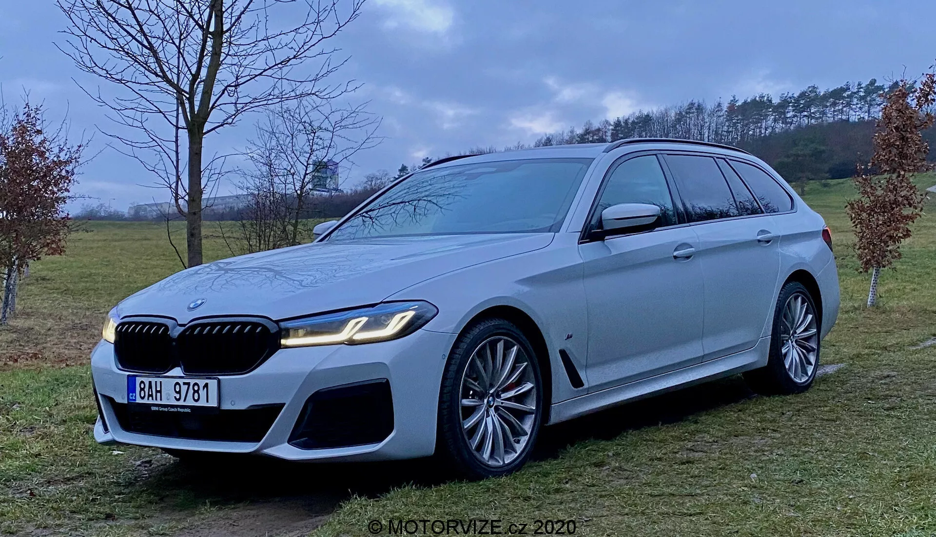 Imagem de uma perua de luxo BMW Série 5 Touring 2020 (BMW 520i Touring Steptronic) na cor prata clara ou cinza, exibindo sua dianteira agressiva e esportiva com grandes entradas de ar, grade proeminente e elegantes faróis de LED. O veículo possui grandes rodas de liga leve com design de múltiplos raios e pinças de freio notáveis, uma longa linha de teto com spoiler, acabamentos em cromo nas janelas e uma linha de ombro forte. Fotografado a partir da altura média do veículo em um ambiente semi-rural gramado com iluminação difusa.
