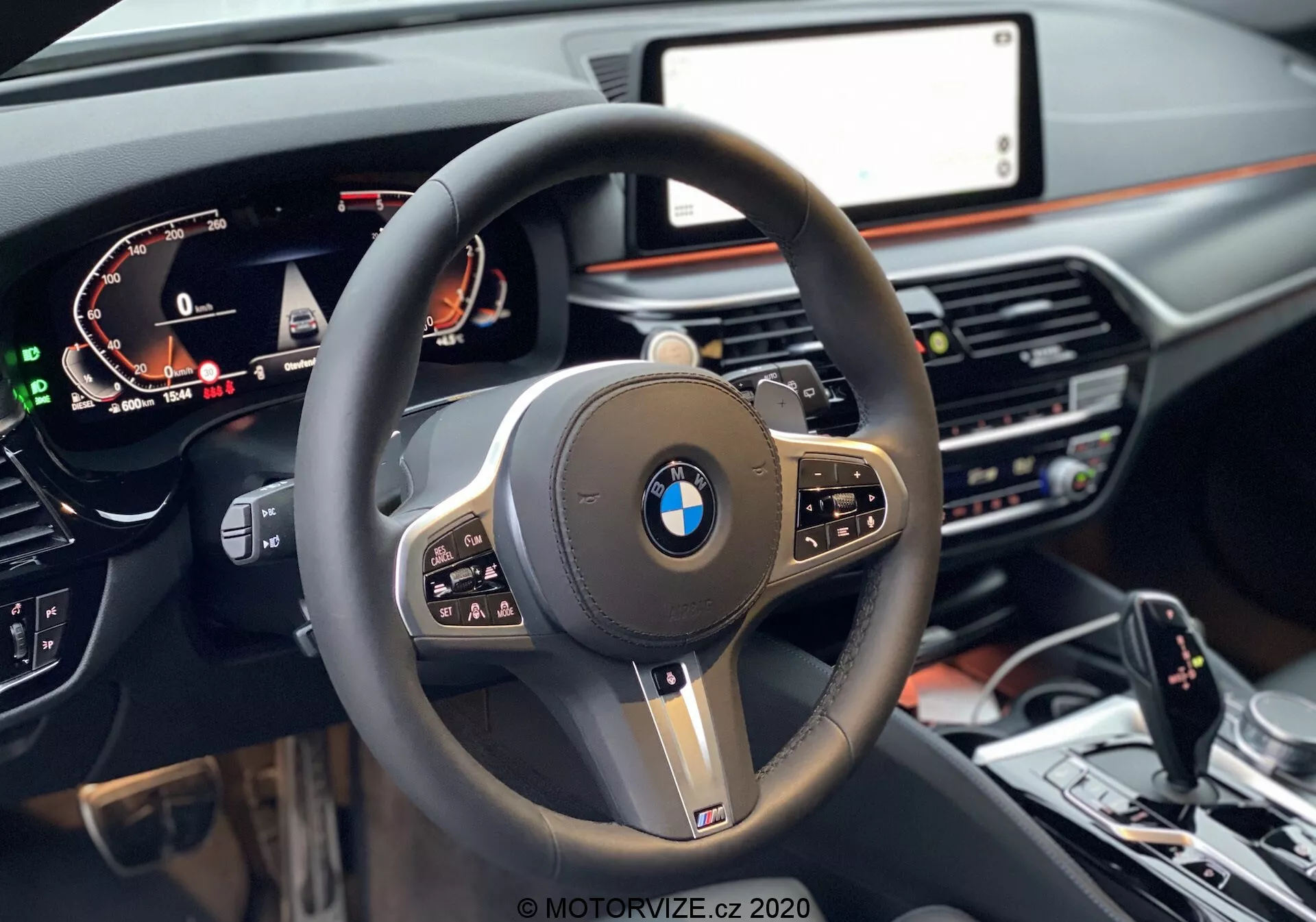 Vista em primeira pessoa do assento do motorista de um BMW 520i Touring Steptronic 2020, com foco no volante moderno de três raios com aro grosso revestido em couro, botões multifuncionais e um logotipo BMW proeminente no centro, rodeado por acabamento preto brilhante e acentos metálicos. Um painel de instrumentos digital à esquerda exibe a velocidade, RPM e informações do motorista, enquanto um display central de infotainment é parcialmente visível à direita. O painel de tons escuros, com um toque de acabamento em madeira, possui controles de climatização e sistema de mídia abaixo da tela. Na parte inferior da imagem, vê-se o console central com o seletor de marchas, controlador iDrive e botões de controle de dinâmicas de condução.