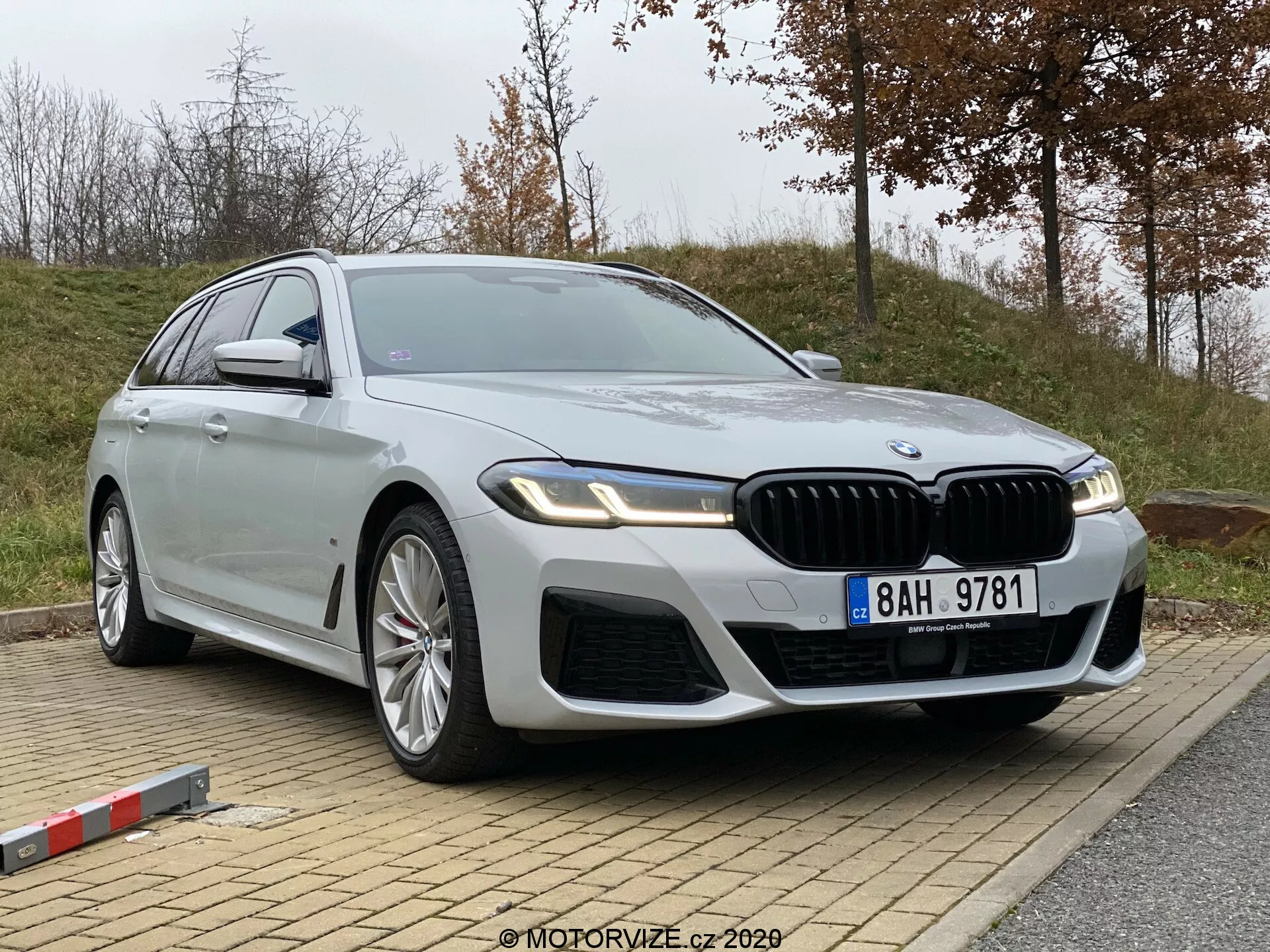 A imagem mostra o BMW Série 5 2020 (520i Touring Steptronic), um sedã de luxo moderno, estacionado em uma leve inclinação. Capturada de um ângulo baixo e ligeiramente de lado frontal, destaca o quarto dianteiro e o perfil do veículo. Os detalhes notáveis incluem uma grade em forma de rim proeminente, entradas de ar intricadas no para-choques dianteiro, faróis de LED angulares elegantes, grandes rodas de liga leve multirraios, uma silhueta de corpo longo e fluida com acabamento prata metálico, acabamento cromado, sinais de mudança de direção integrados nos espelhos laterais e recursos tecnológicos avançados potenciais, como assistentes de condução autônoma e assistência de estacionamento. Um emblema é visível no painel do quarto dianteiro, indicando a variante específica do modelo.