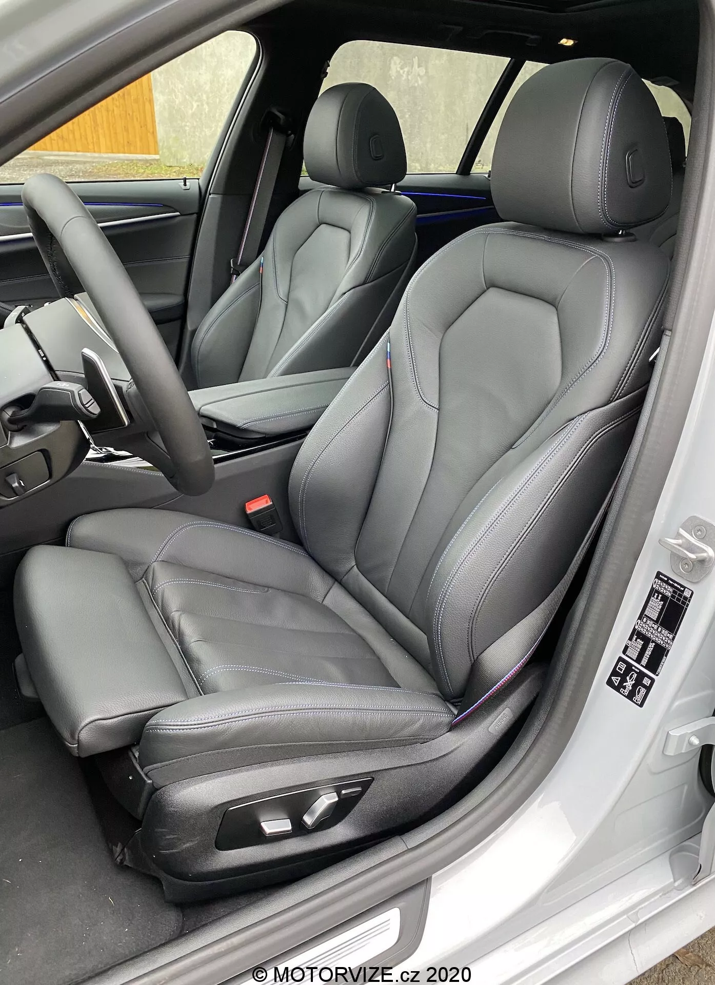 Vista lateral em close-up do assento do motorista do BMW 520i Touring Steptronic 2020, apresentando assentos dianteiros modernos estilo balde em couro preto com costura contrastante, com controles de ajuste eletrônico para maior conforto. A imagem também captura o painel interno da porta do motorista com controles de janelas e espelhos elétricos, configurações de memória do assento e outras características do painel e volante indicativas de luxo e tecnologia avançada.