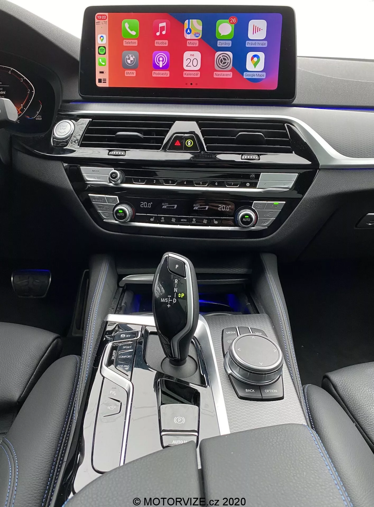 Imagem do interior de um BMW 520i Touring 2020 com transmissão Steptronic, tirada da perspectiva do motorista e ligeiramente elevada para dar uma visão mais clara do console central, área da alavanca de câmbio e parte do painel. A imagem apresenta uma grande tela de infotainment exibindo uma interface de conexão de smartphone, provavelmente Apple CarPlay, montada no alto do painel para fácil visibilidade. Abaixo da tela estão as saídas de ar centrais modernas e horizontais com detalhes cromados, seguidas por botões táteis e mostradores para controle climático. O console central também abriga os controles do sistema de áudio, controles climáticos adicionais e um possível slot para CD player. A alavanca de câmbio ergonômica e minimalista, provavelmente eletrônica, é cercada por acabamentos em preto brilhante e metálico, com botões de seleção do modo de condução e um botão rotativo para navegação. O interior também inclui apoios de braço estofados em couro com costura visível, acrescentando um toque de luxo. Iluminação branca suave destaca os botões ao redor da área da alavanca de câmbio, indicando uma transmissão automática.