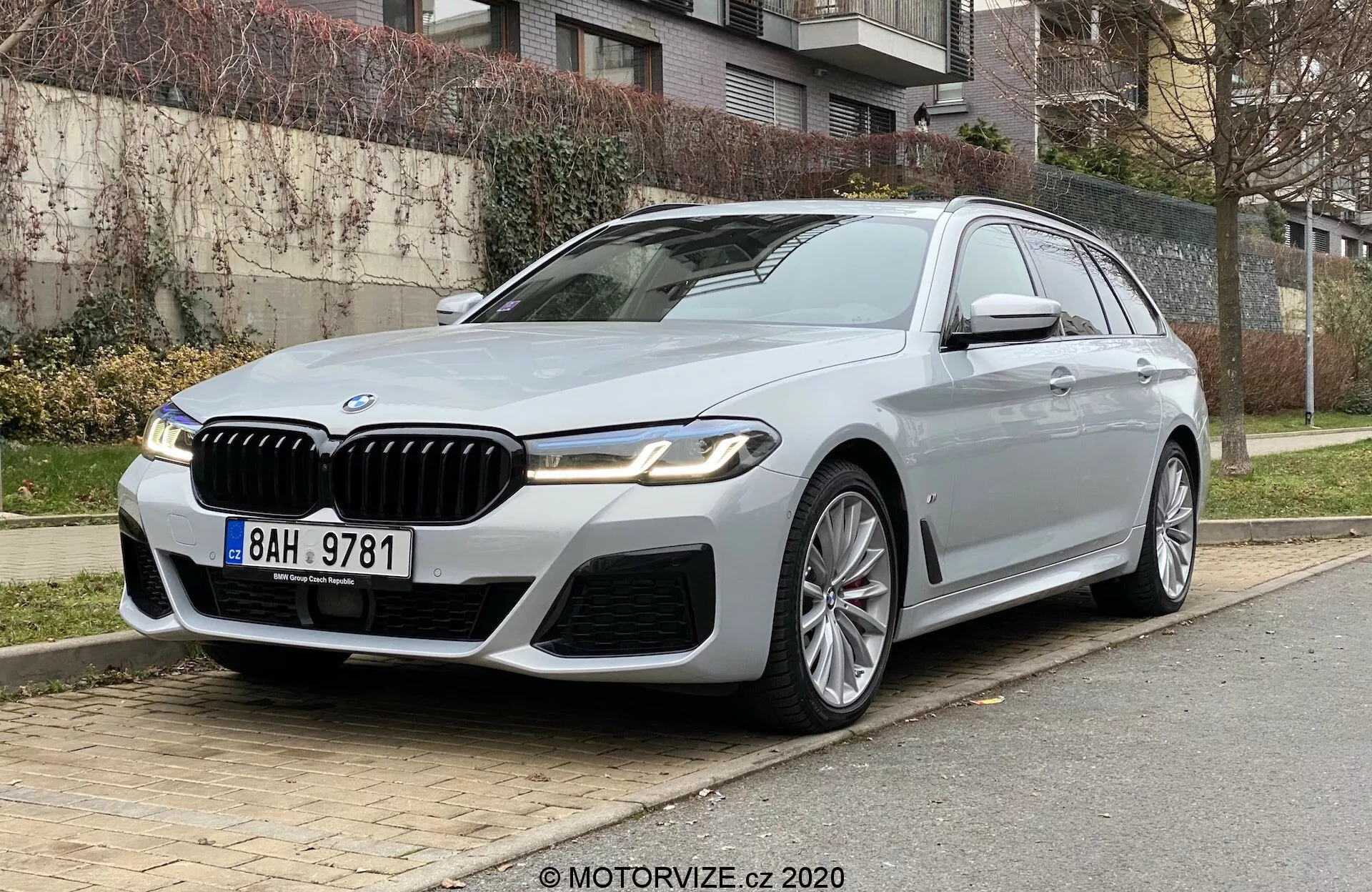 Luxuoso BMW 520i Touring estacionado em uma rua da cidade, visto de um ângulo frontal baixo de três quartos que enfatiza a frente e o lado do motorista. O carro possui uma grade de rim proeminente com detalhes cromados, faróis de LED elegantes, para-choque dianteiro esportivo e vincos definidos no capô. Rodas de liga leve com múltiplos raios, espelhos laterais com indicadores de direção e câmera, moldura cromada das janelas e teto solar acrescentam ao visual luxuoso. Posicionado do lado direito com grama, arbustos e edifícios residenciais ao fundo, sugerindo tecnologia avançada e design de alto padrão.