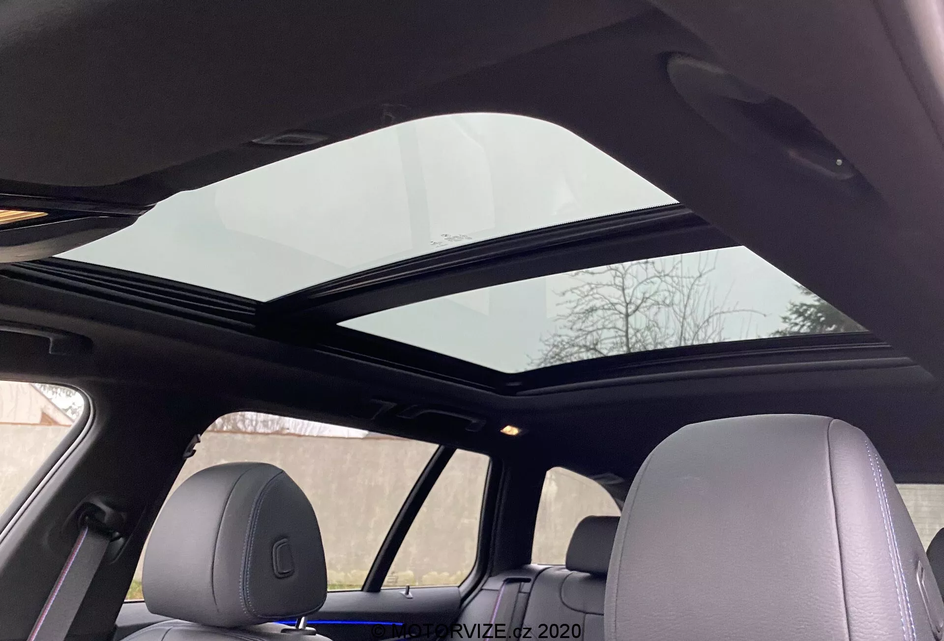 Vista interior de um BMW Série 5 2020 520i Touring com transmissão Steptronic exibindo um teto solar panorâmico que se estende sobre os bancos dianteiros e traseiros a partir da perspectiva de um passageiro no banco traseiro olhando para cima. O painel frontal do teto solar está ligeiramente inclinado aberto, melhorando a ventilação. O revestimento do teto é de cor clara, complementando o design interior coerente do veículo, com os apoios de cabeça combinando com o material interno, destacando o conforto avançado e as comodidades modernas.