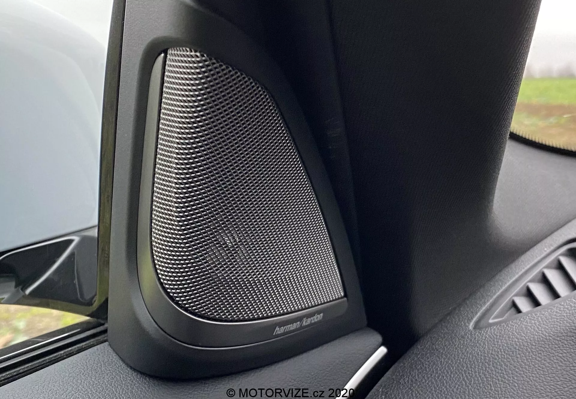 Visão de perto do interior do BMW 5er-Reihe 2020 (BMW 520i Touring Steptronic), focando no alto-falante premium da porta da Harman Kardon. A imagem captura o padrão de malha fina da grade metálica do alto-falante, cercada por materiais plásticos pretos ou de toque suave, projetados para amortecer vibrações e oferecer qualidade de som superior. O ângulo da câmera mostra a parte superior do painel da porta, com o alto-falante em destaque e uma pequena parte do apoio de braço da porta. Um leve desfoque do fundo destaca esse recurso de áudio de alta qualidade neste veículo de luxo.