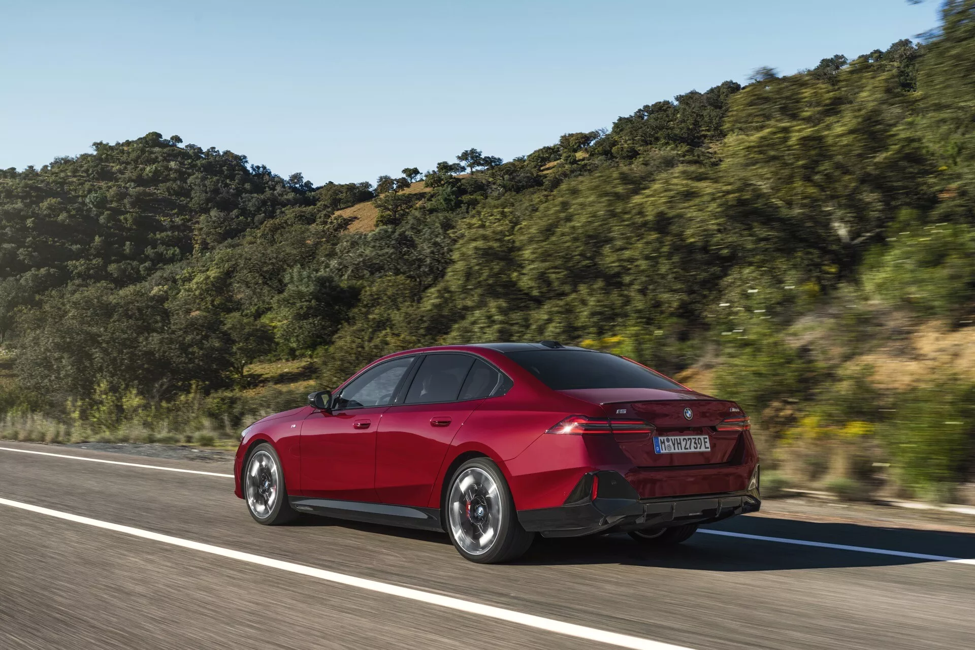 Sedã vermelho de quatro portas BMW Série 5 (G60-G61) em movimento capturado por um ângulo traseiro de três quartos, destacando elementos de design traseiro e lateral como a forma do porta-malas, para-choque traseiro, lanternas traseiras LED e linhas aerodinâmicas contra um fundo montanhoso sob céu claro