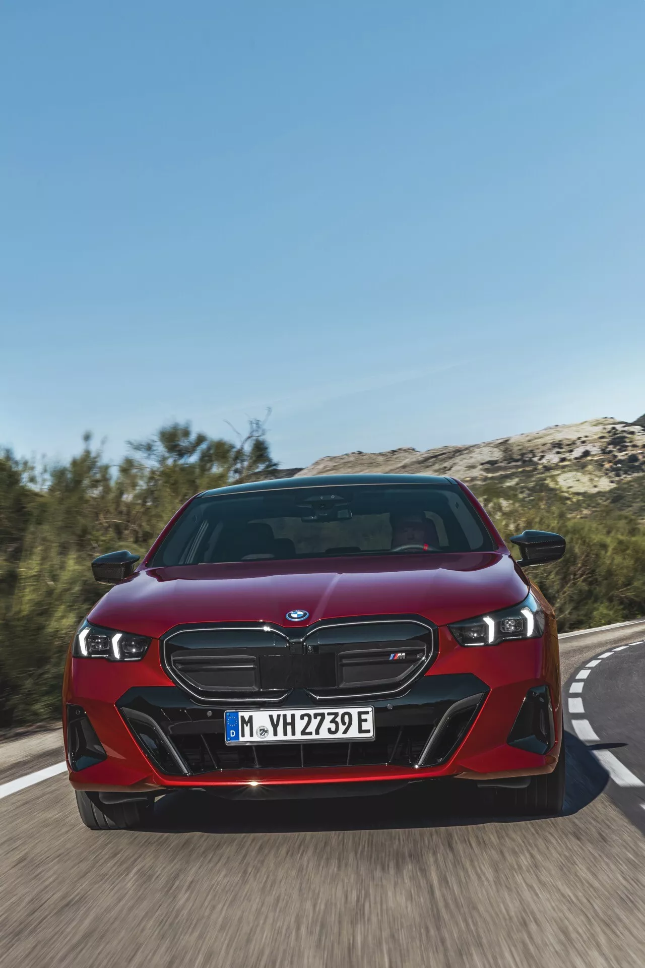 BMW Série 5 (G60-G61) 2023 em vermelho, exibindo sua grade dianteira dupla de rins distintiva, faróis LED e fascia frontal dinâmica de um ângulo frontal baixo em uma estrada, destacando a velocidade do veículo e os recursos avançados eletrificados iPerformance
