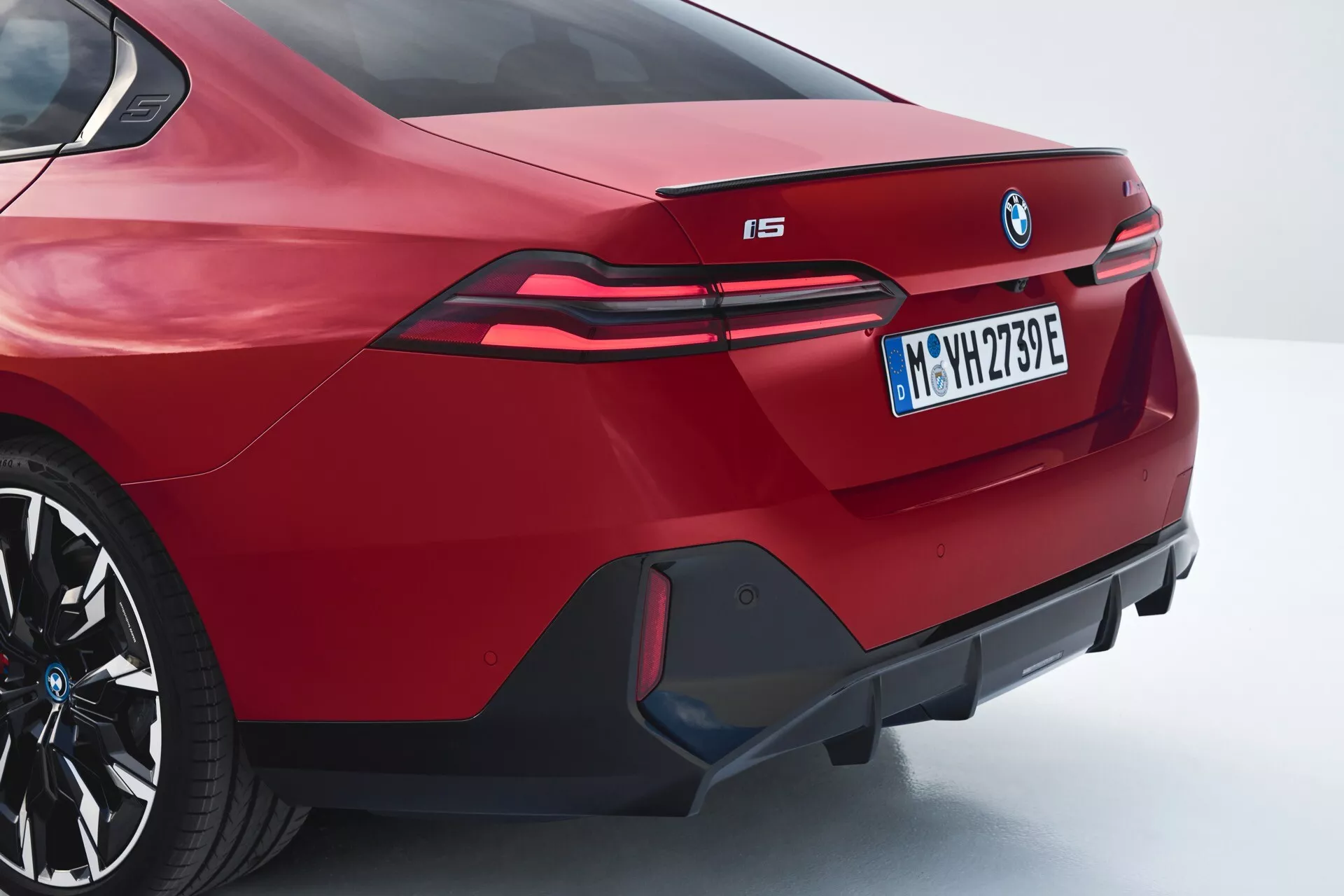 Vista traseira angular de um BMW i5 2023 vermelho mostrando o design esportivo do sedan, spoiler integrado, emblema BMW proeminente, lanternas traseiras LED alongadas estendendo-se para os para-lamas traseiros, designação do modelo elétrico 'i5', para-choque traseiro agressivo com difusor decorativo e saídas de exaustão falsas, em perfil traseiro e lateral de três quartos.