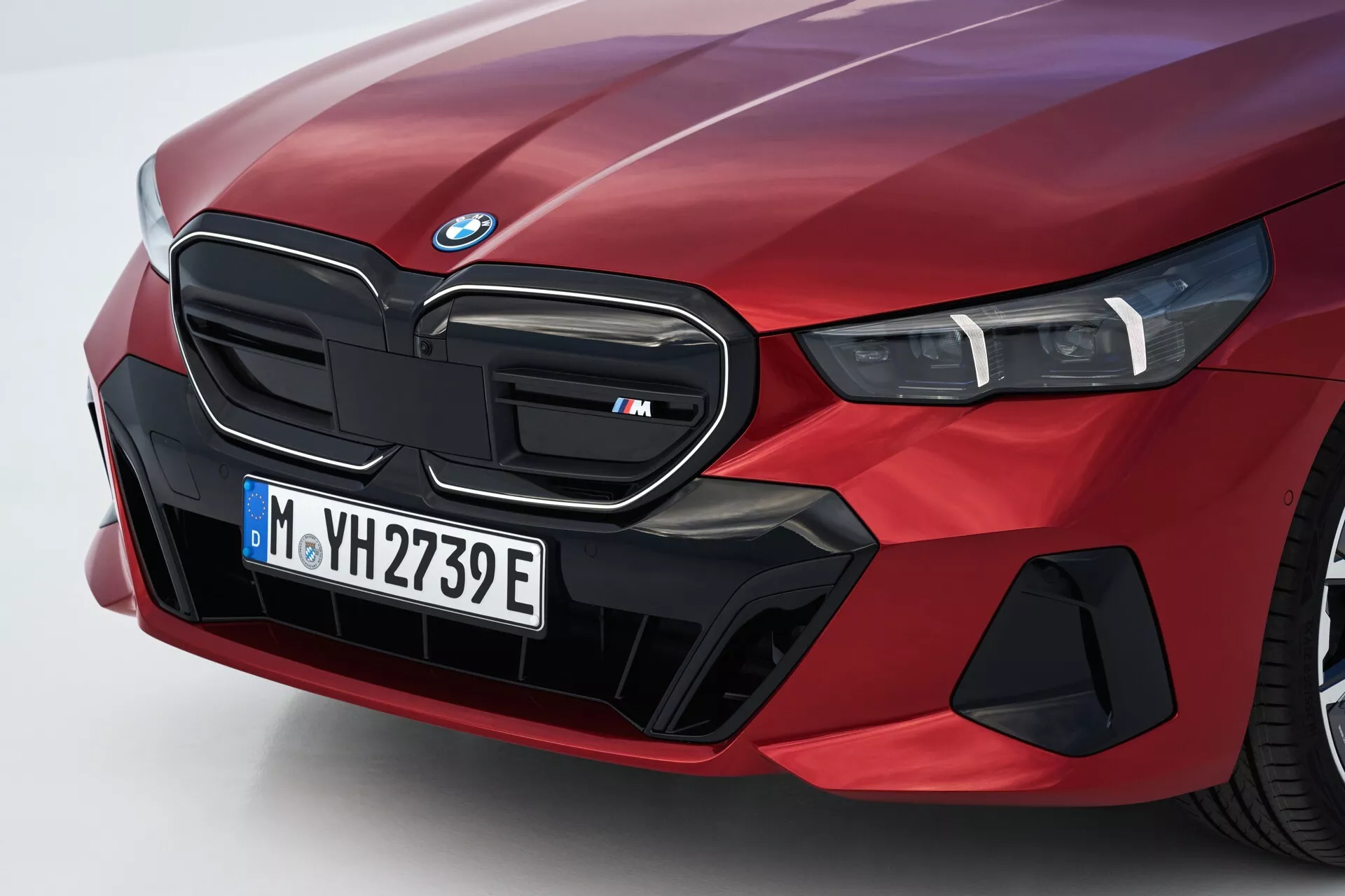 Vista angular e próxima do lado frontal esquerdo de um BMW Série 5 (G60-G61) 2023 em vermelho, apresentando a grelha renal icônica marcada como modelo 'M', conjunto de faróis em LED e elementos aerodinâmicos especializados sugerindo capacidades elétricas ou híbridas