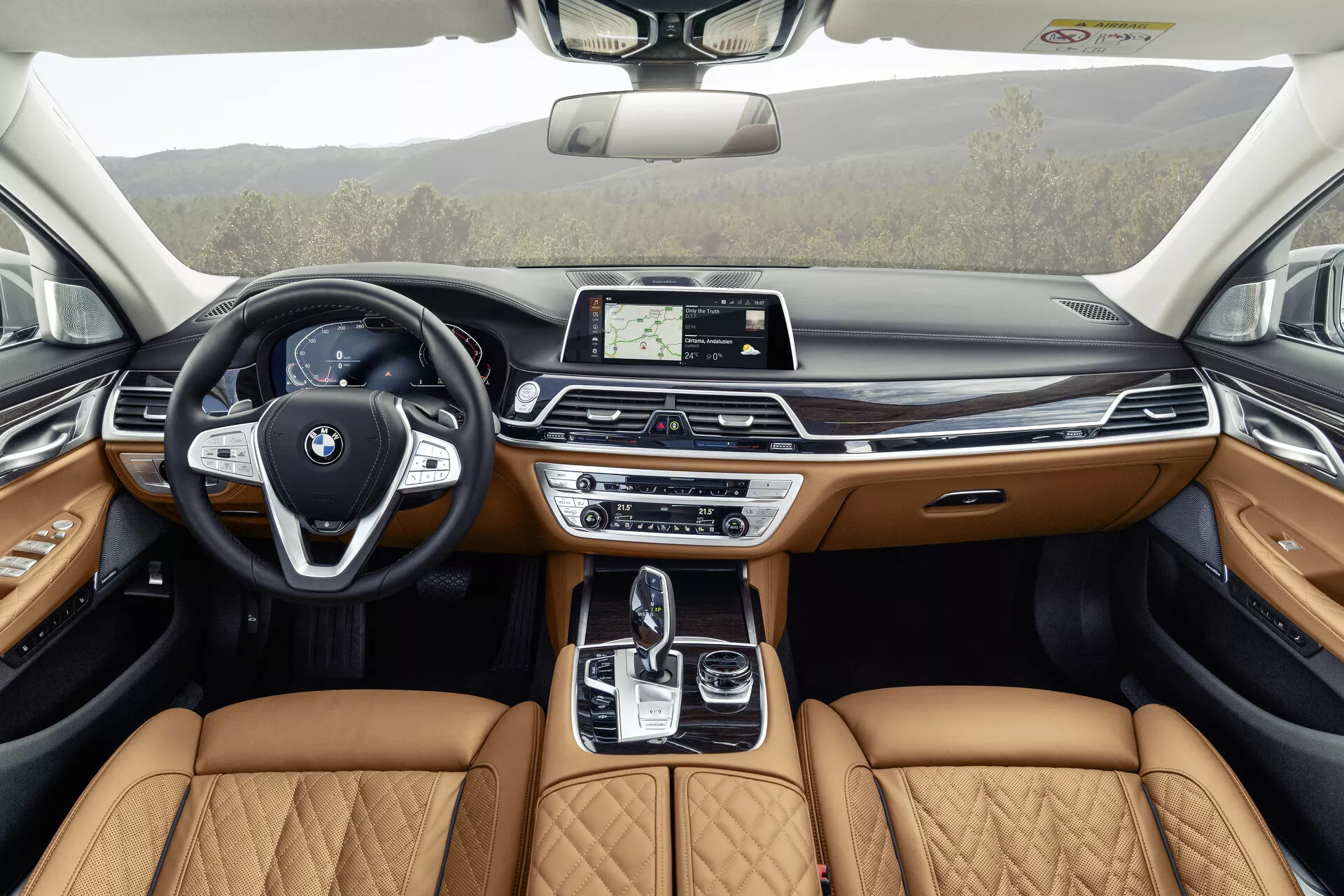 A imagem mostra o interior de um BMW Série 7 2019 (facelift G11/G12) desde a perspetiva do banco traseiro, destacando o design detalhado e luxuoso da cabine dianteira. Com um moderno volante com botões multifuncionais e um emblema BMW proeminente, o painel de instrumentos elegante aloja um ecrã tátil central para infotainment. A consola central inclui uma alavanca de mudanças automática, um painel tátil e vários sistemas de controlo dinâmico, complementados por assentos de couro bege acolchoados que indicam um nível de acabamento premium. Os painéis das portas refletem este luxo com couro bege, acabamentos em madeira e iluminação ambiente, sugerindo que funcionalidades avançadas de tecnologia e conforto estão integradas em todo o veículo, incluindo um teto solar para luz natural adicional.