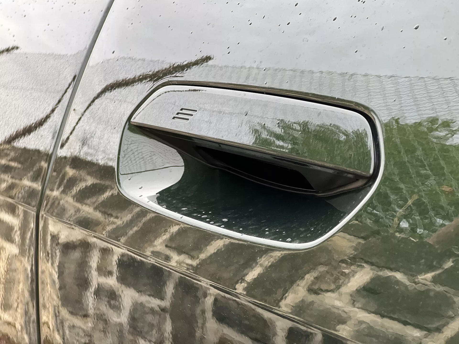 Vista detalhada do espelho lateral do lado do motorista do BMW i4 2021 mostrando seu design aerodinâmico, caixa preta com uma fina textura de linha horizontal, acabamento prateado, superfície resistente à água com gotas de chuva, e sua característica dobrável refletida no vidro salpicado pela chuva.