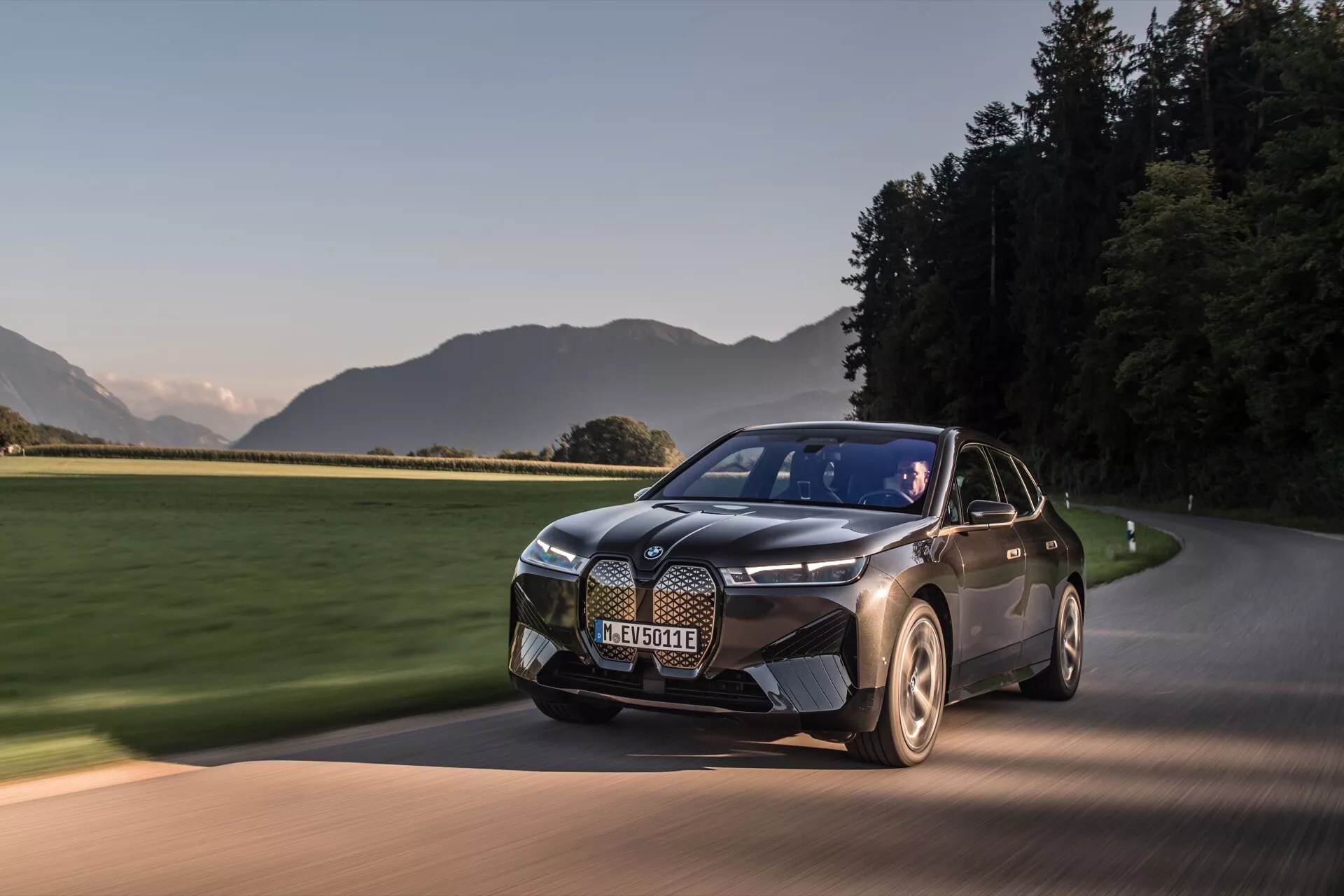 SUV elétrico moderno BMW iX 2021 (i20) em movimento em uma estrada pavimentada ao amanhecer ou ao entardecer, mostrando sua pintura em dois tons, design elegante com uma grade proeminente, faróis distintos e rodas aerodinâmicas contra um cenário cênico.