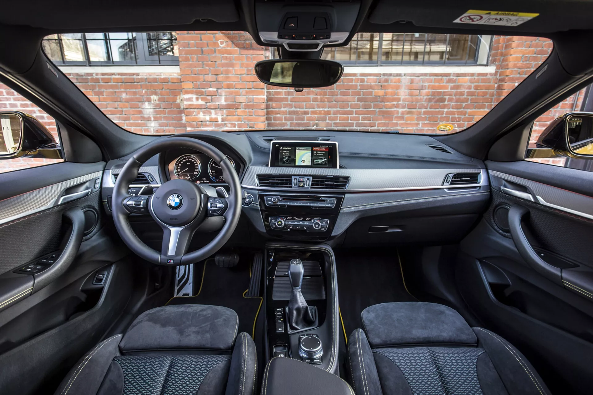 Vista interior de um BMW X2 2018, mostrando a cabine frontal a partir da área de assentos traseiros, destacando o volante com controles montados e logotipo da BMW, cluster de instrumentos digital-analógico, console central com alavanca de câmbio e sistema iDrive, display de infotainment não touch, painel de instrumentos luxuoso com acabamento em madeira ou metal, controle de clima de zona dupla, assentos dianteiros contornados, porta-copos práticos e compartimentos de armazenamento, e painéis de porta com recursos de controle, sob um teto solar iluminado pelo sol.