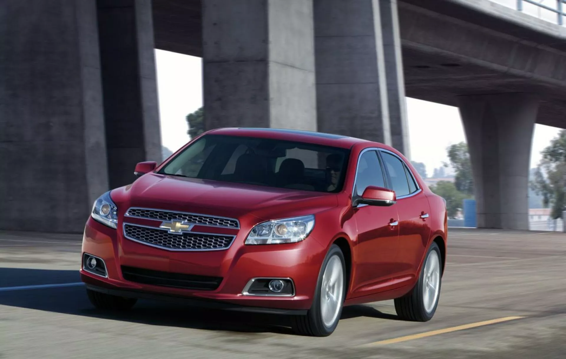 Vista dinâmica de um sedan Chevrolet Malibu 2012 vermelho em movimento em uma ponte, mostrando o design frontal com grade cromada, faróis elegantes, silhueta esportiva e rodas de liga.