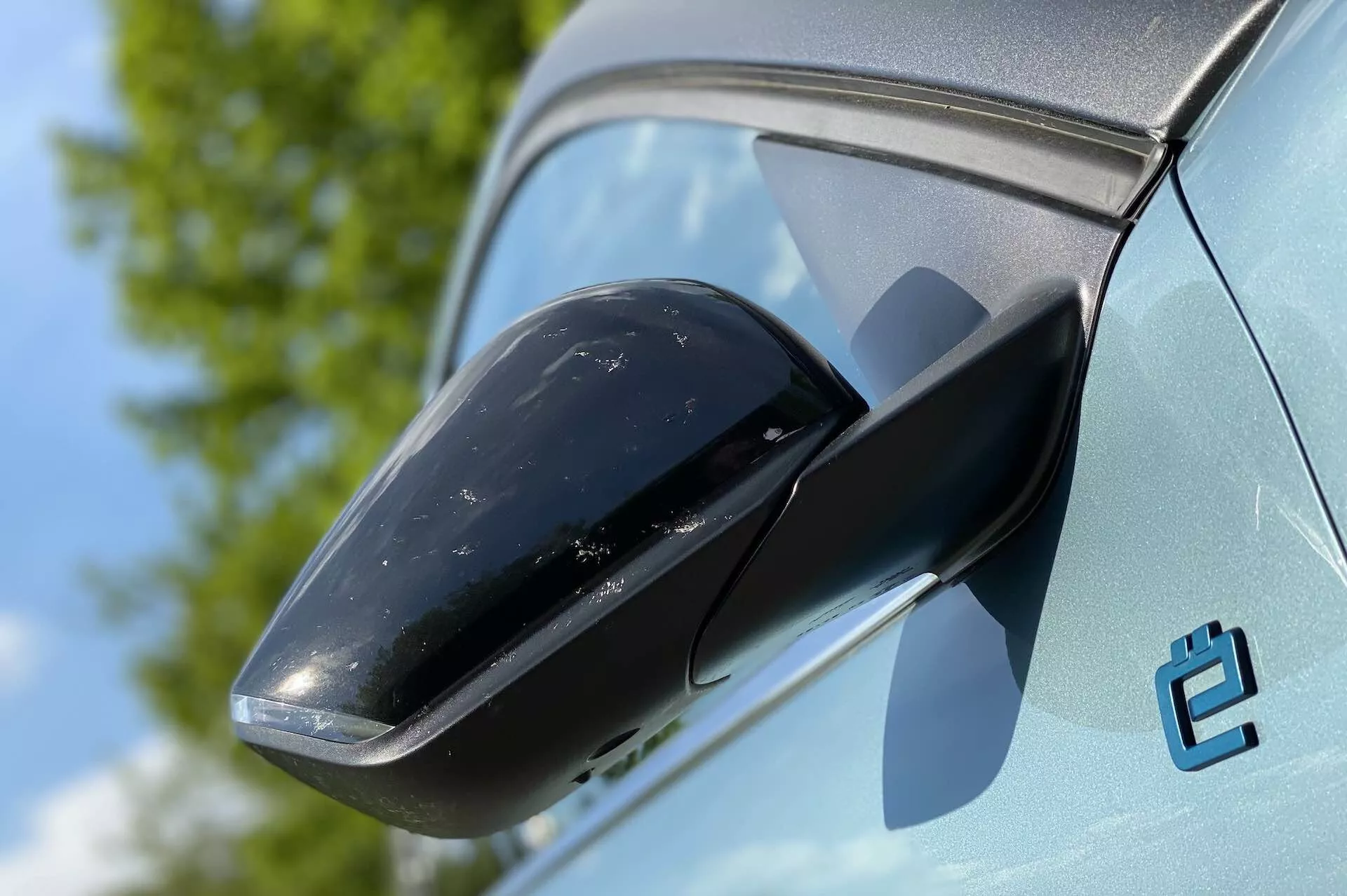 Close-up de um retrovisor lateral do Citroën C4 2020 (E-C4), enfatizando a carcaça preta do espelho e a cor clara da carroceria. O fundo revela um céu claro e vegetação, sugerindo um ambiente externo. A condição do carro é nova, embora haja um pouco de sujeira na carcaça do espelho. Um emblema estilizado 'e' também é visível, possivelmente indicando uma variante elétrica.