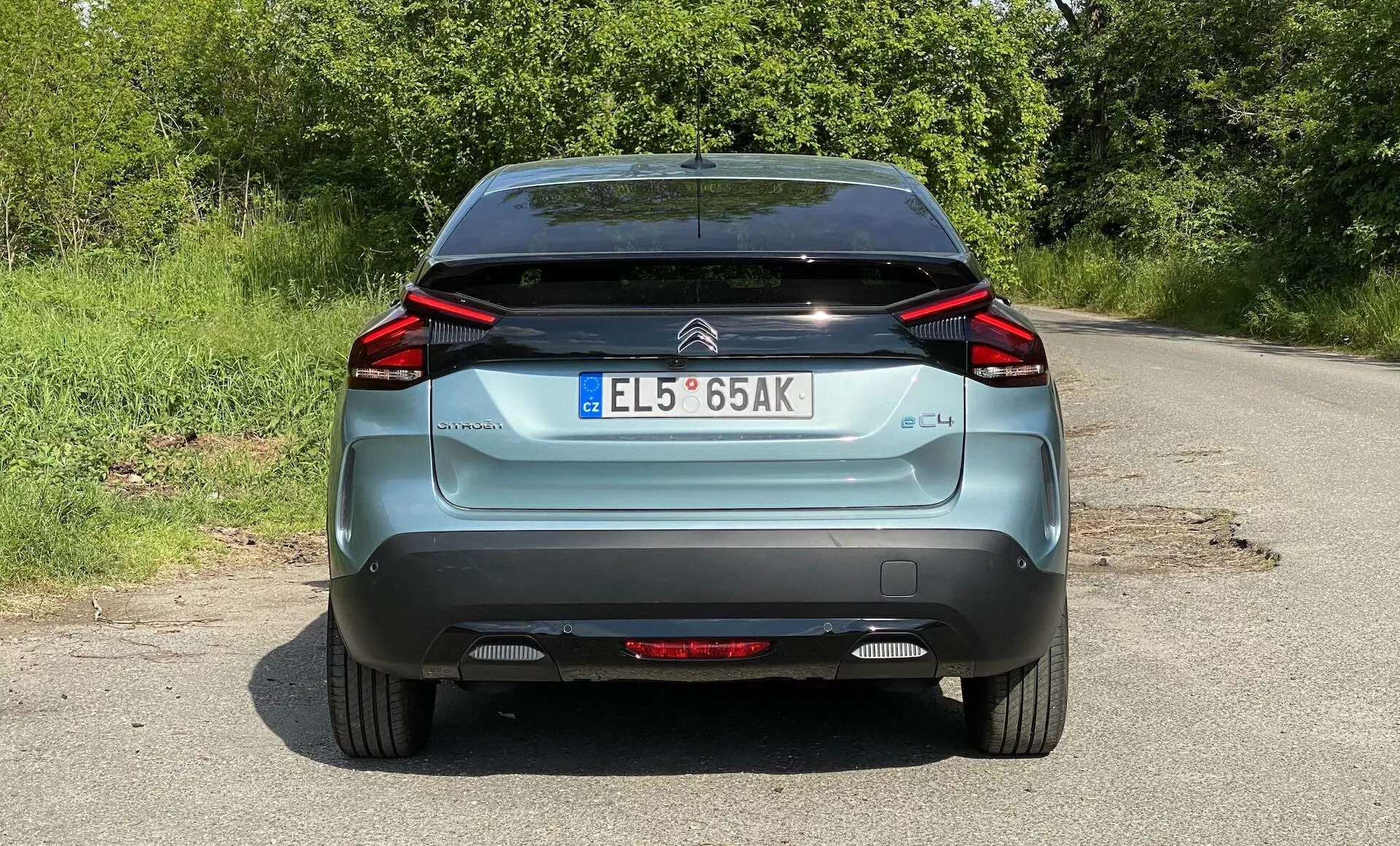 Vista traseira de um Citroën C4 azul de 2020 (e-C4), estacionado em uma estrada rural. A imagem captura toda a largura do veículo, da parte inferior das rodas ao teto, mostrando os característicos faróis traseiros de LED angulares, o emblema da marca, a etiqueta 'C4', a placa de matrícula no estilo europeu, o limpador do para-brisa traseiro e a seção inferior do para-choque preta. Também são visíveis os refletores e a luz de nevoeiro traseira.