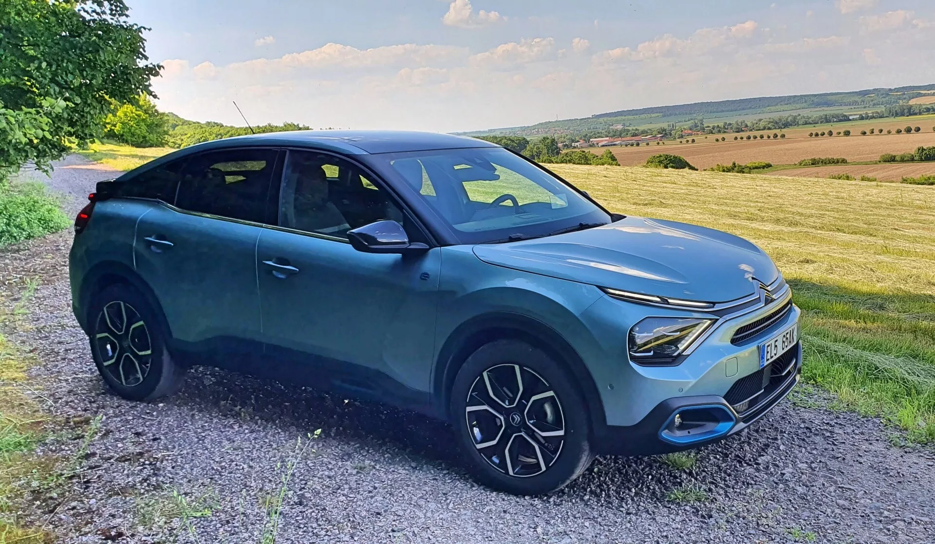 A imagem mostra uma vista frontal lateral do Citroen C4 2020 (e-C4), estacionado em um caminho de cascalho ou beira de estrada com um cenário rural pitoresco com suaves colinas e um céu espaçoso. O crossover SUV moderno tem altura aumentada, proteções ao redor dos arcos das rodas, faróis de LED distintivos, grade minimalista, rodas multi-raios elegantes e uma carroceria azul suave acentuada por elementos pretos. O design é elegante e aerodinâmico com uma postura esportiva, indicando categorias como SUVs, Carros Modernos, Veículos Crossover, Design de Veículos, Tecnologia Automotiva e potencialmente Veículos Elétricos.