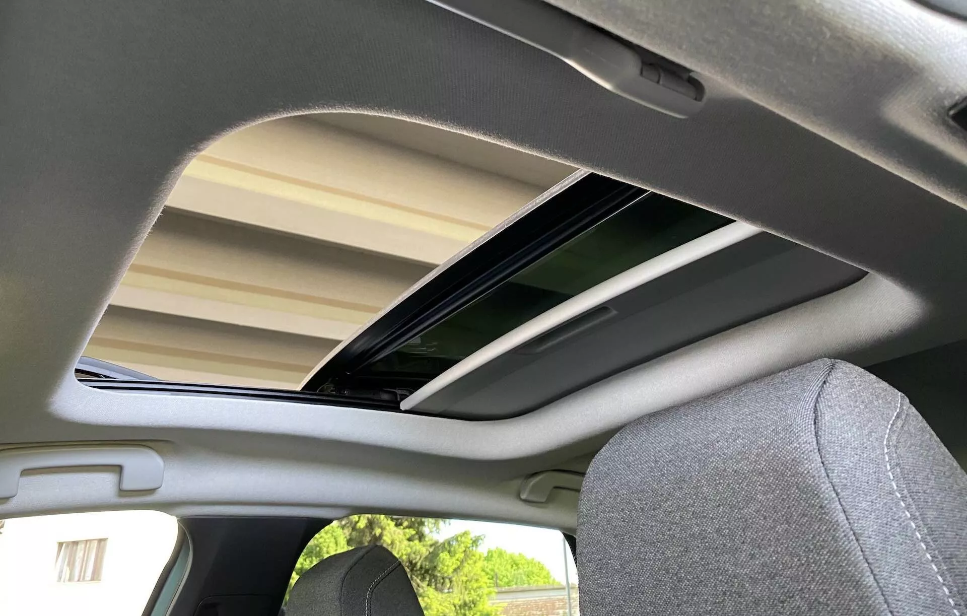 Vista interior do banco traseiro de um Citroën C4 2020 (e-C4), focada no teto solar aberto a partir de um ângulo baixo, ligeiramente fora do centro para a esquerda. O teto solar de vidro está inclinado e aberto, permitindo que a luz natural ilumine o forro de tecido cinza claro ou bege e o encosto de cabeça do assento do passageiro correspondente. A imagem captura a atmosfera arejada e os detalhes essenciais do design interior do carro.