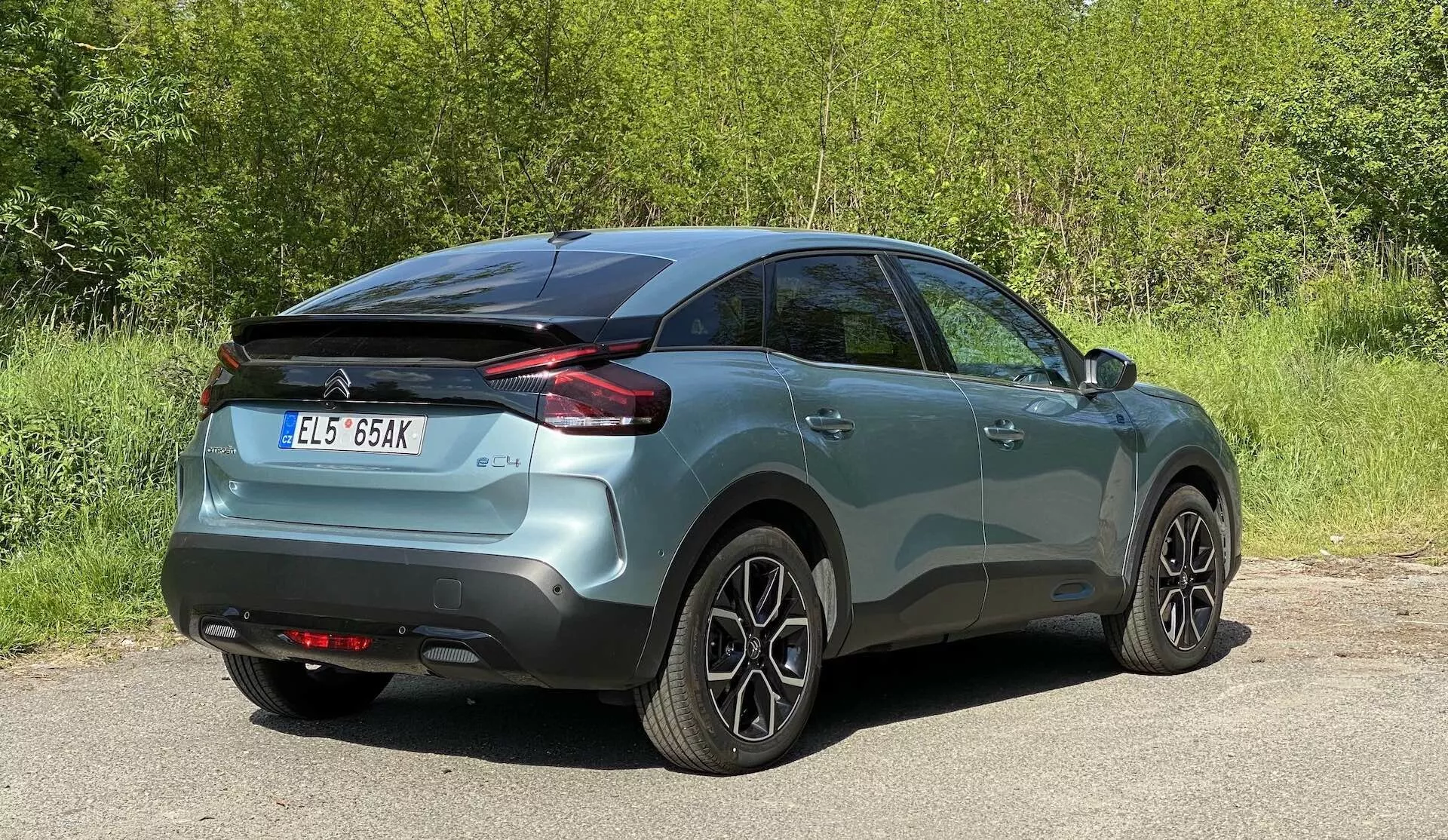 Imagem de um carro Citroen C4 2020 (e-C4) mostrando o ângulo traseiro de três quartos. O veículo pintado em metal está centralmente posicionado, revelando seus perfis laterais e traseiros, incluindo luzes traseiras de design horizontal, maçanetas das portas da cor da carroceria e rodas de liga leve multiraiadas. O ambiente sugere um ambiente ao ar livre com alguma vegetação ao fundo.