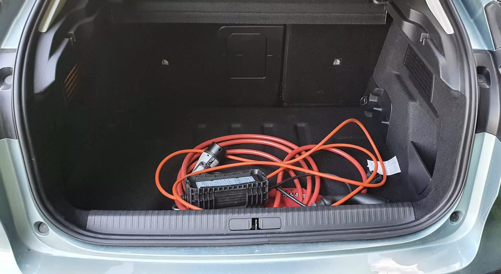 Vista de cima para baixo do porta-malas do Citroen C4 2020 (e-C4) hatchback, mostrando o piso acarpetado, painéis laterais com um painel de acesso e uma extensão laranja colocada de maneira solta conectada a um inversor de energia portátil. O limiar do porta-malas apresenta uma proteção de borracha ou plástico, destacando o design prático de armazenamento do veículo.