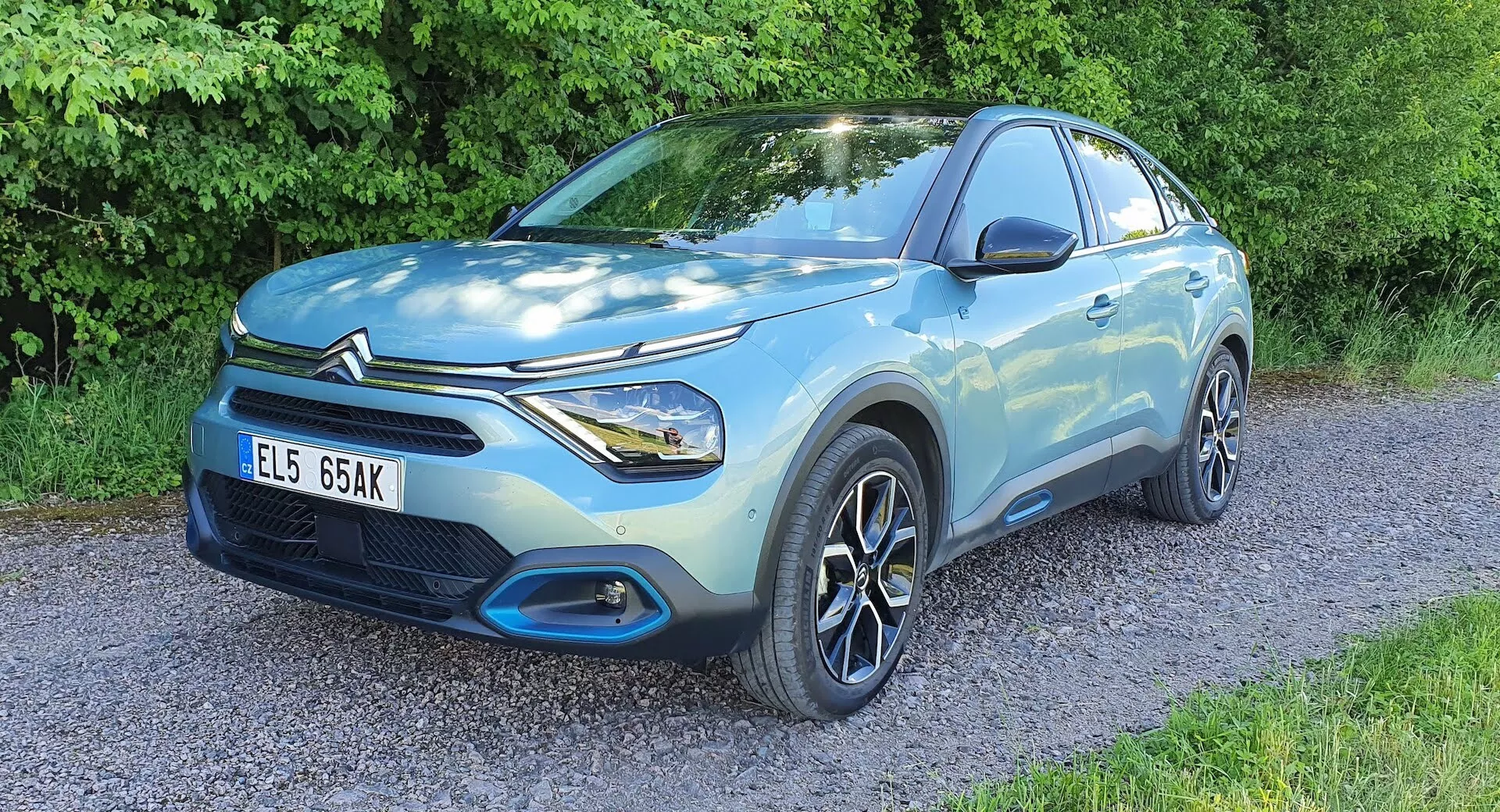 Citroen C4 2020 (e-C4) SUV compacto azul claro estacionado em superfície de cascalho com vegetação ao fundo, visão frontal de três quartos mostrando o lado do motorista, a fachada frontal, carroceria esculpida, faróis de LED, rodas de liga leve multirraios, grade frontal e emblema do fabricante, destacando características de design e tecnologia.