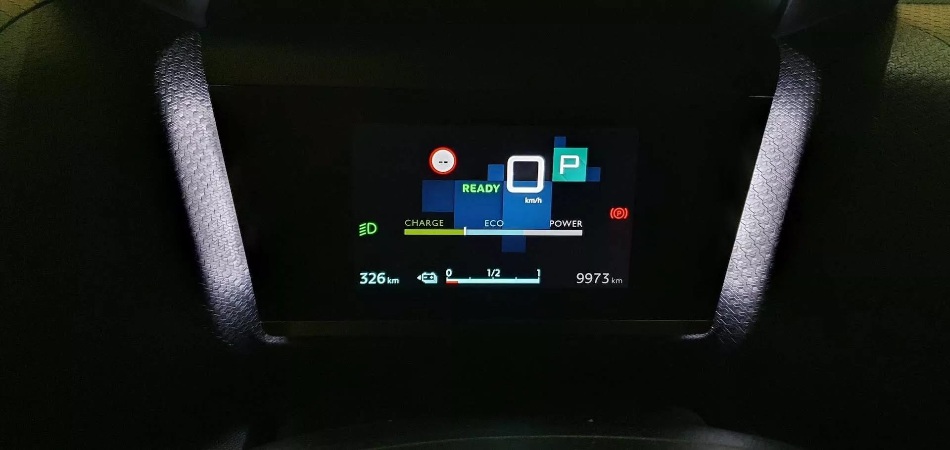 Vista aproximada do painel de instrumentos digital do Citroen C4 2020 (e-C4), mostrando indicadores 'CHARGE' e 'ECO', sinal 'READY', ícone de bateria com '32.6', ícone de folha ecológica, indicador de uso de 'POWER', símbolo de estacionamento 'P', '10' com seta para baixo indicando temperatura e leitura do odômetro de '9973 km', capturada do ponto de vista do assento do motorista.