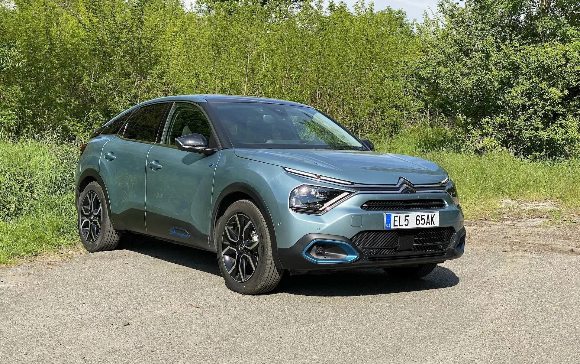 Uma visão completa de um Citroën C4 2020 (e-C4) estacionado ao ar livre, tirada de um ângulo frontal de três quartos mostrando a grade frontal, o design elegante dos faróis de LED e a forma do capô, juntamente com um perfil lateral parcial incluindo portas e espelhos laterais. O hatchback com elementos de design crossover/SUV apresenta rodas de liga leve modernas, sinais de direção integrados nos espelhos e um acabamento de pintura metálica. O fundo é de vegetação e o carro é mostrado à luz do dia natural sem elementos distrativos.