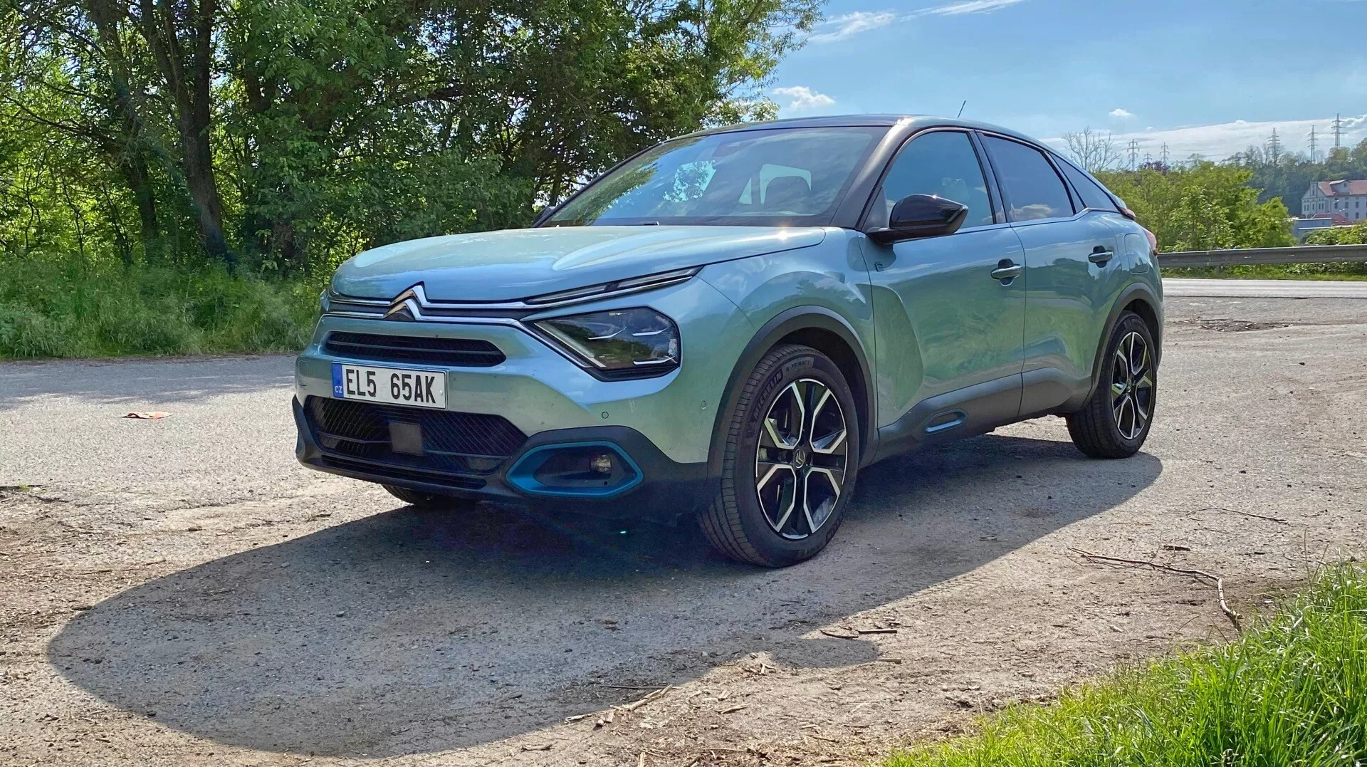 A imagem mostra um Citroen C4 (e-C4) hatchback ou SUV compacto de 2020 de um ângulo de três quartos frontal baixo. Esta perspectiva destaca a frente, incluindo a grade, faróis e para-choques, bem como o perfil lateral do motorista, apresentando o para-lama frontal, a roda, a porta do motorista e parte da porta traseira. O carro tem um design moderno e esportivo com linhas suaves, um teto inclinado, faróis dianteiros LED distintos e uma grade dianteira proeminente. O para-choque dianteiro tem um acabamento preto contrastante, realçando sua aparência robusta. O design intrincado das rodas com vários raios sugere um foco em estilo ou desempenho. A pintura metálica brilha intensamente ao sol, refletindo um exterior limpo e polido. O veículo está estacionado ao ar livre em uma paisagem verde, indicada pelas árvores e condições climáticas claras. Características tecnológicas como sensores ou câmeras não são confirmadas a partir deste ângulo, e a placa está visível, mas omitida por questões de privacidade.