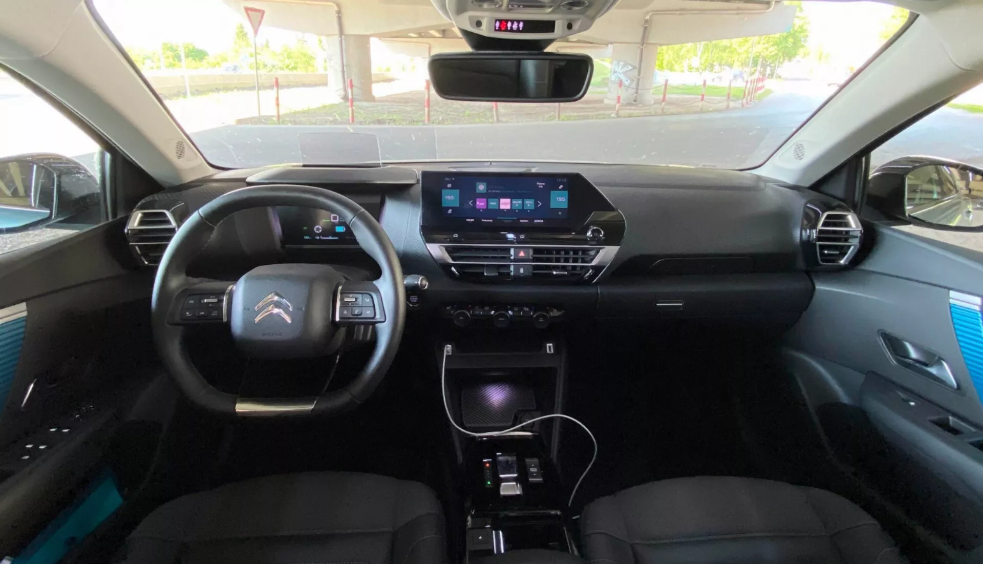 Vista interna do Citroen C4 2020 (e-C4) do banco traseiro, focalizando o painel com um cluster digital, volante moderno com controles integrados e uma tela de infotainment proeminente no console central. O console abriga botões de controle de clima, alavanca de câmbio e controles adicionais, sugerindo recursos como navegação GPS, sistema de multimídia e controle de clima manual ou automático. Os bancos dianteiros parecem estofados com encostos de cabeça e os painéis das portas exibem detalhes premium.