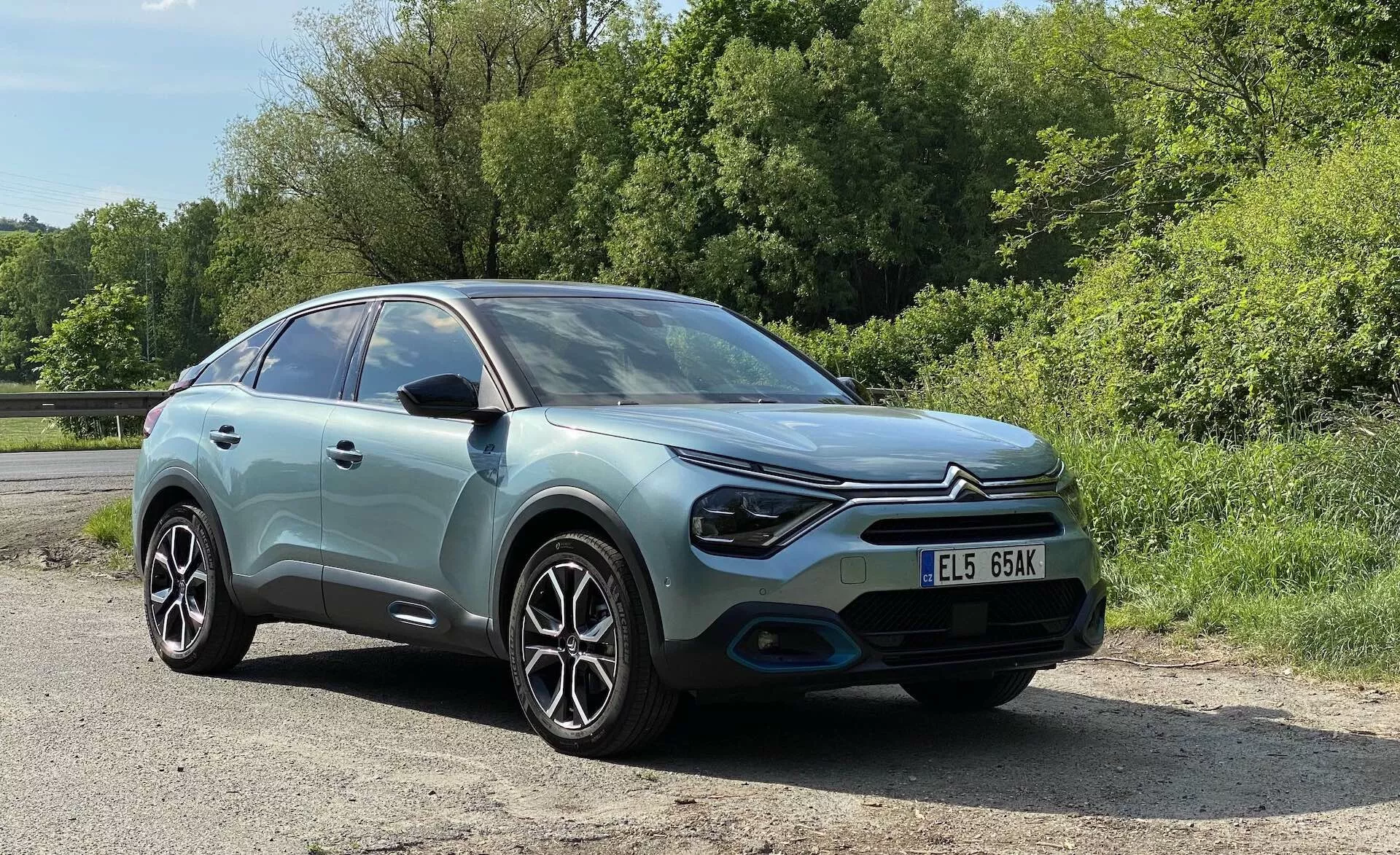 Imagem de um Citroen C4 2020 (e-C4) capturada de uma perspetiva frontal de três quartos, destacando o seu corpo azul pastel com um contraste no teto preto, faróis LED modernos, para-choques dianteiro esculpido, rodas de liga leve e um padrão de raios intricado. A imagem, tirada de um ligeiro ângulo para baixo, proporciona vistas detalhadas do farol do lado esquerdo, do para-choques dianteiro, da roda dianteira, do para-brisas e da janela do lado do condutor, com a placa de matrícula claramente visível. O carro está situado em um cenário rural ou parque com vegetação exuberante ao fundo sob a luz do dia ensolarada.