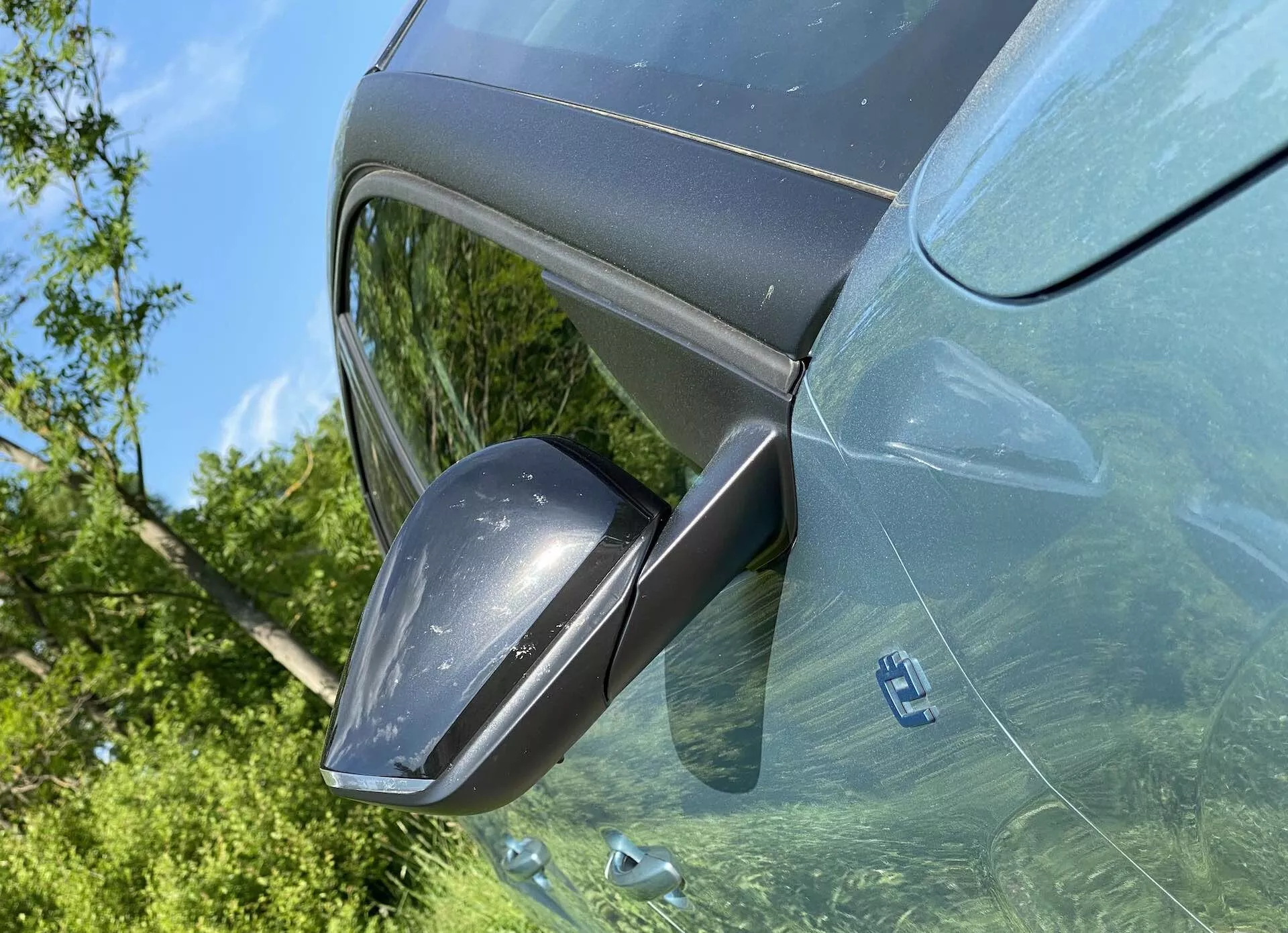 Close-up do lado do motorista do Citroen C4 2020 (e-C4) visto de um ângulo para cima, focando no moderno retrovisor lateral com indicadores de seta integrados, carcaça combinada com a cor do carro e refletindo a vegetação externa nos vidros escurecidos. Uma cena em plena luz do dia destaca a pintura metálica e o design elegante e moderno do veículo.
